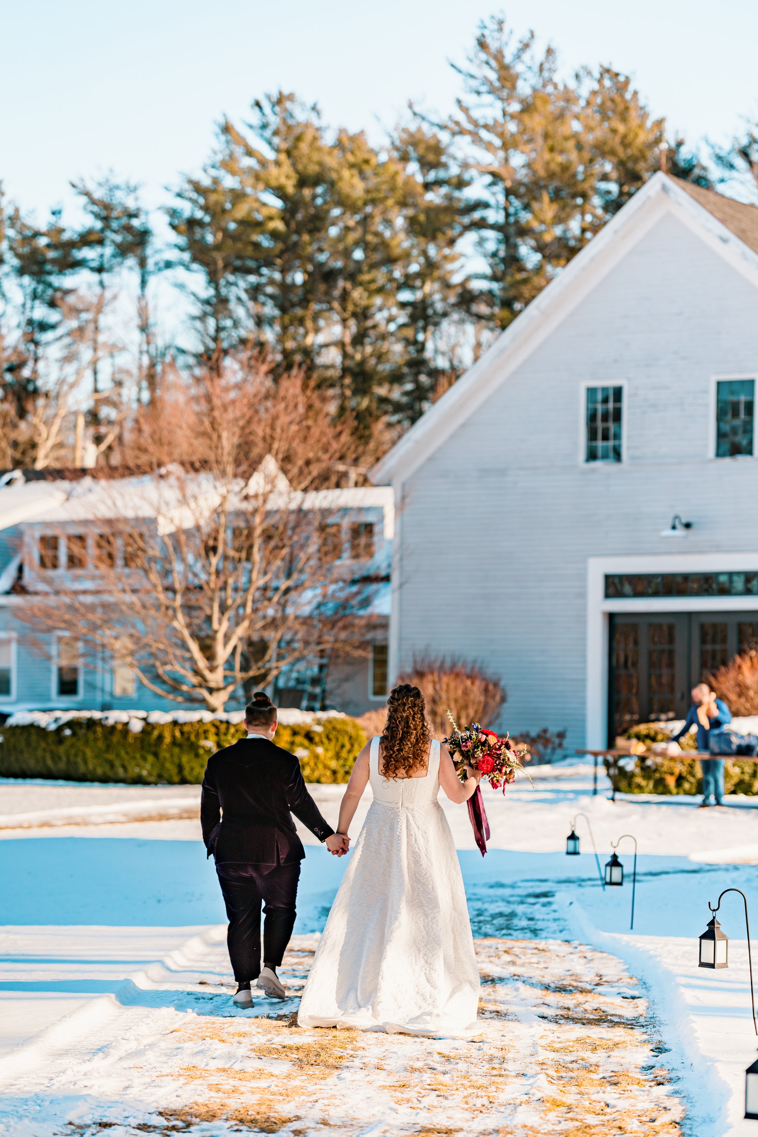 A bride and groom walking hand in hand outside in the snow, with the bride holding a bouquet of flowers, as they head towards a white house.