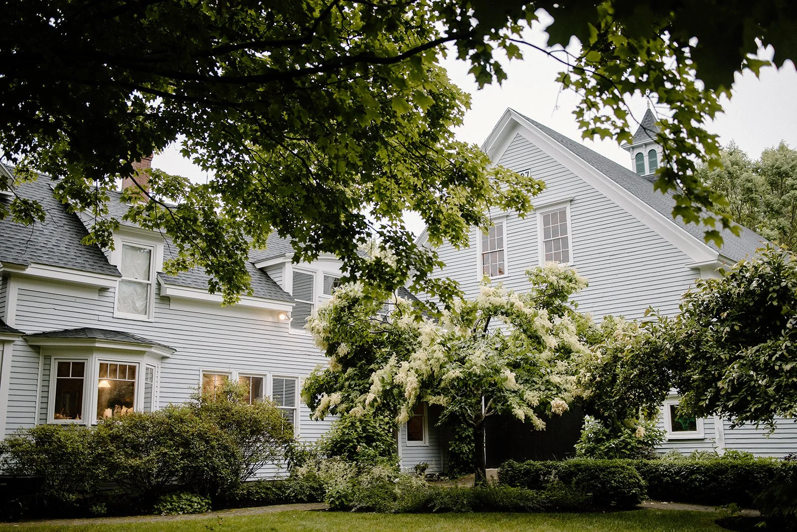 White house with multiple windows surrounded by green trees and bushes, with cloudy sky in the background.
