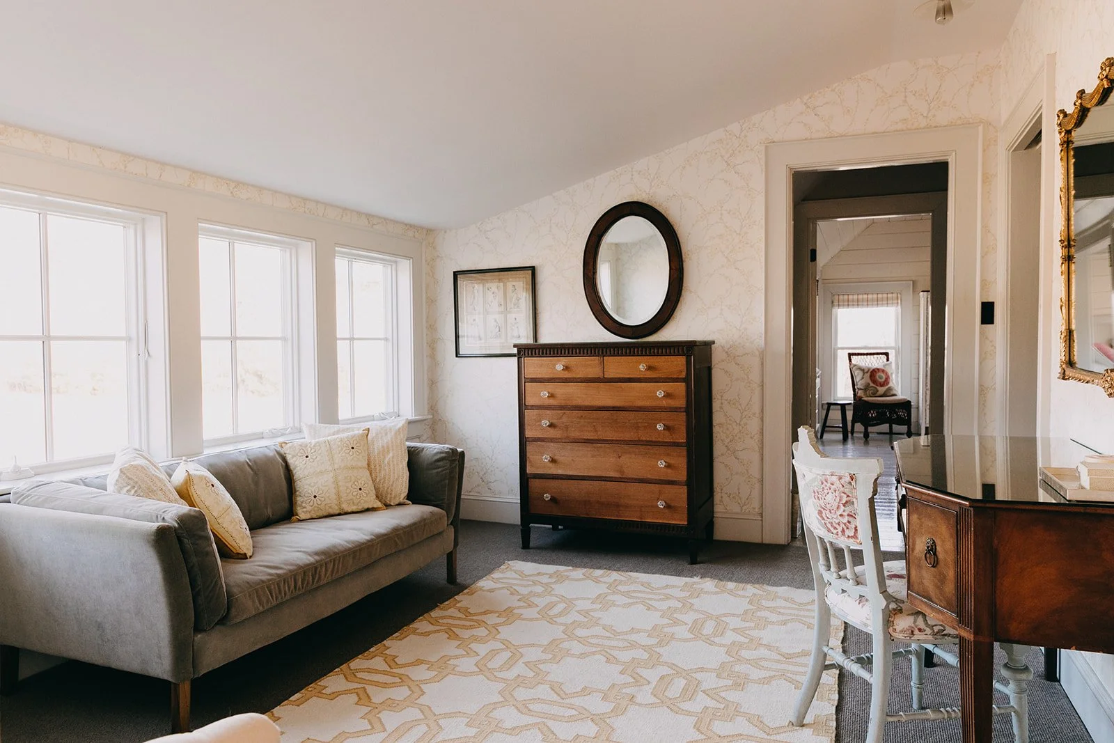 Living room with a gray sofa, beige and patterned pillows, white-framed windows, wooden dresser with a round mirror above, patterned wallpaper, and a doorway leading to a room with a chair.