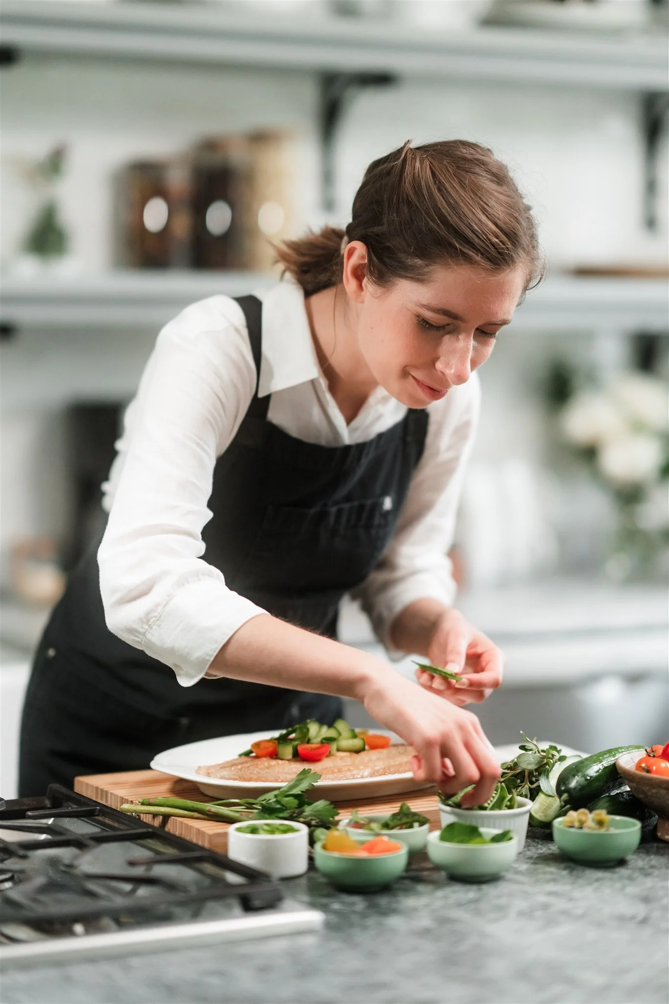 A woman wearing a white shirt and black apron preparing a meal with various fresh vegetables and herbs on a kitchen countertop.