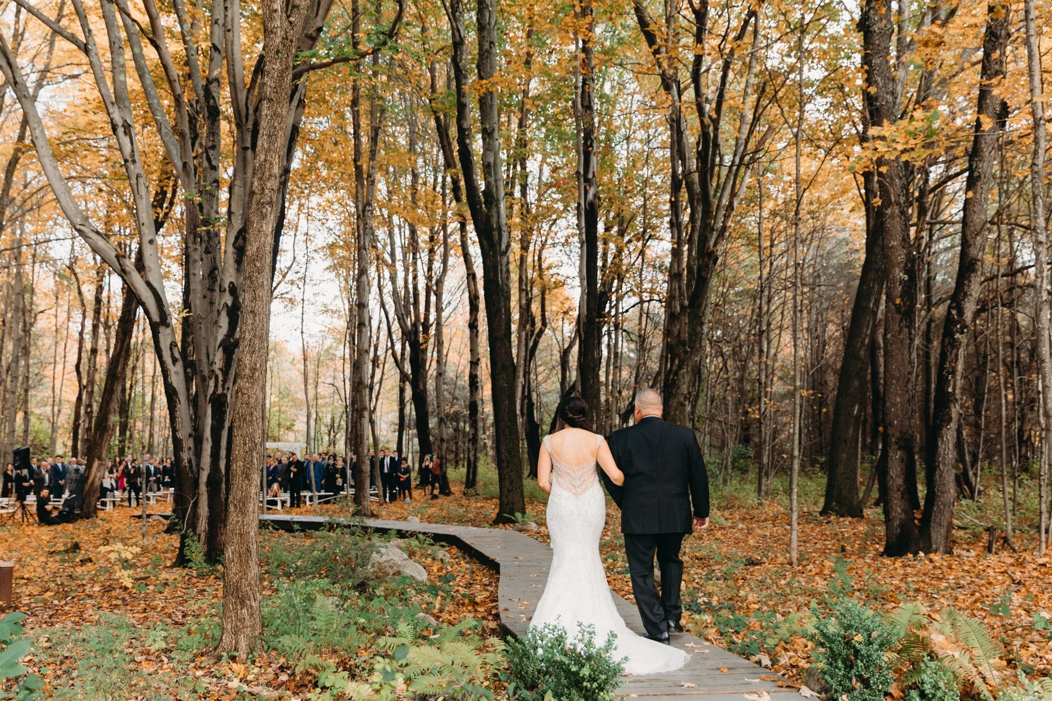 A bride and groom walking arm in arm down a wooded aisle during an outdoor wedding ceremony in fall, with guests seated in the background.