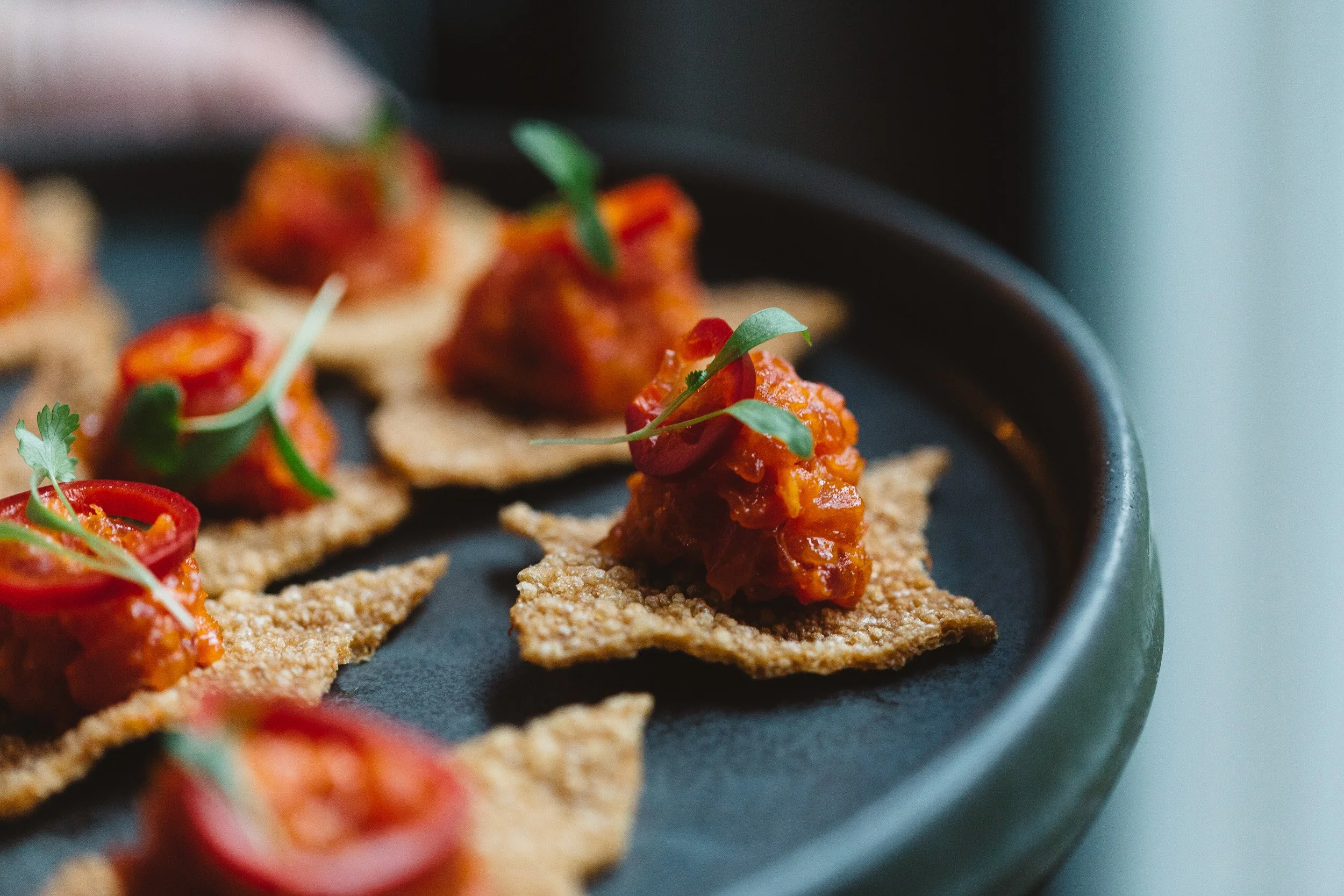 Close-up of small appetizer bites on a black platter, consisting of crispy crackers topped with red chili sauce, cherry tomato slices, and microgreens.