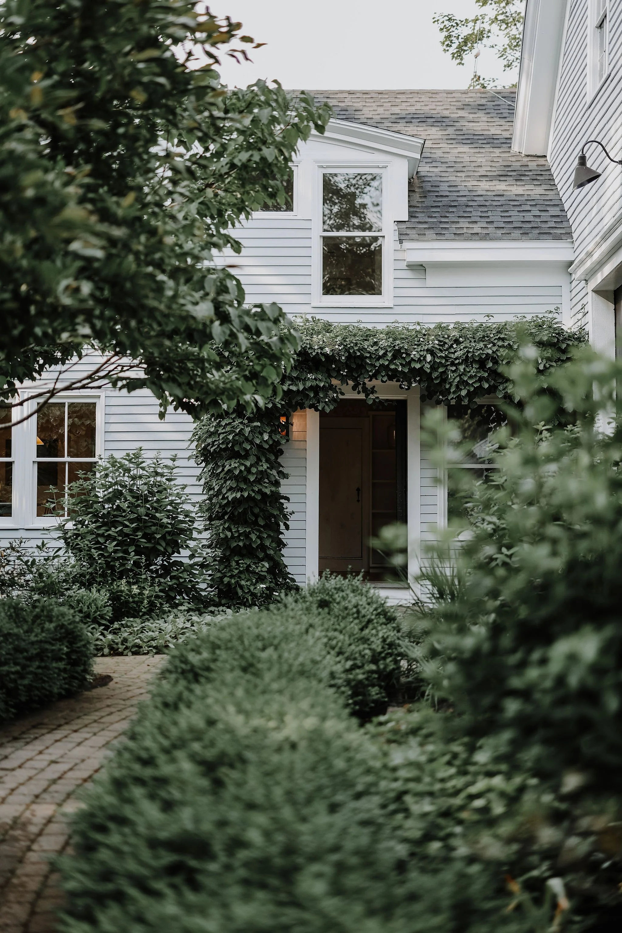 Front view of a white house with a small covered entrance, surrounded by neatly trimmed bushes and greenery, with a brick pathway leading to the door.
