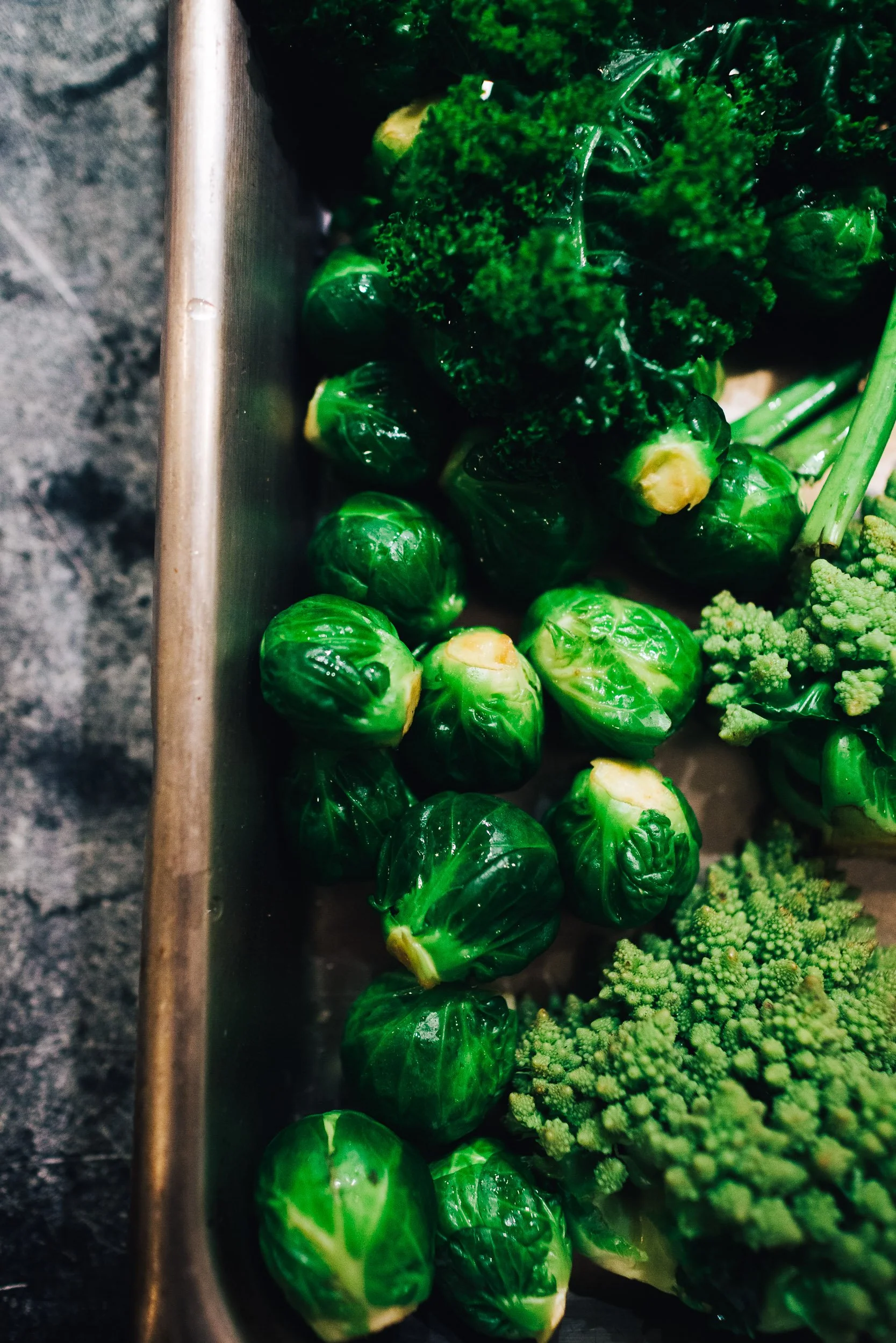Close-up of fresh Brussels sprouts, broccoli, and cauliflower in a stainless steel container.