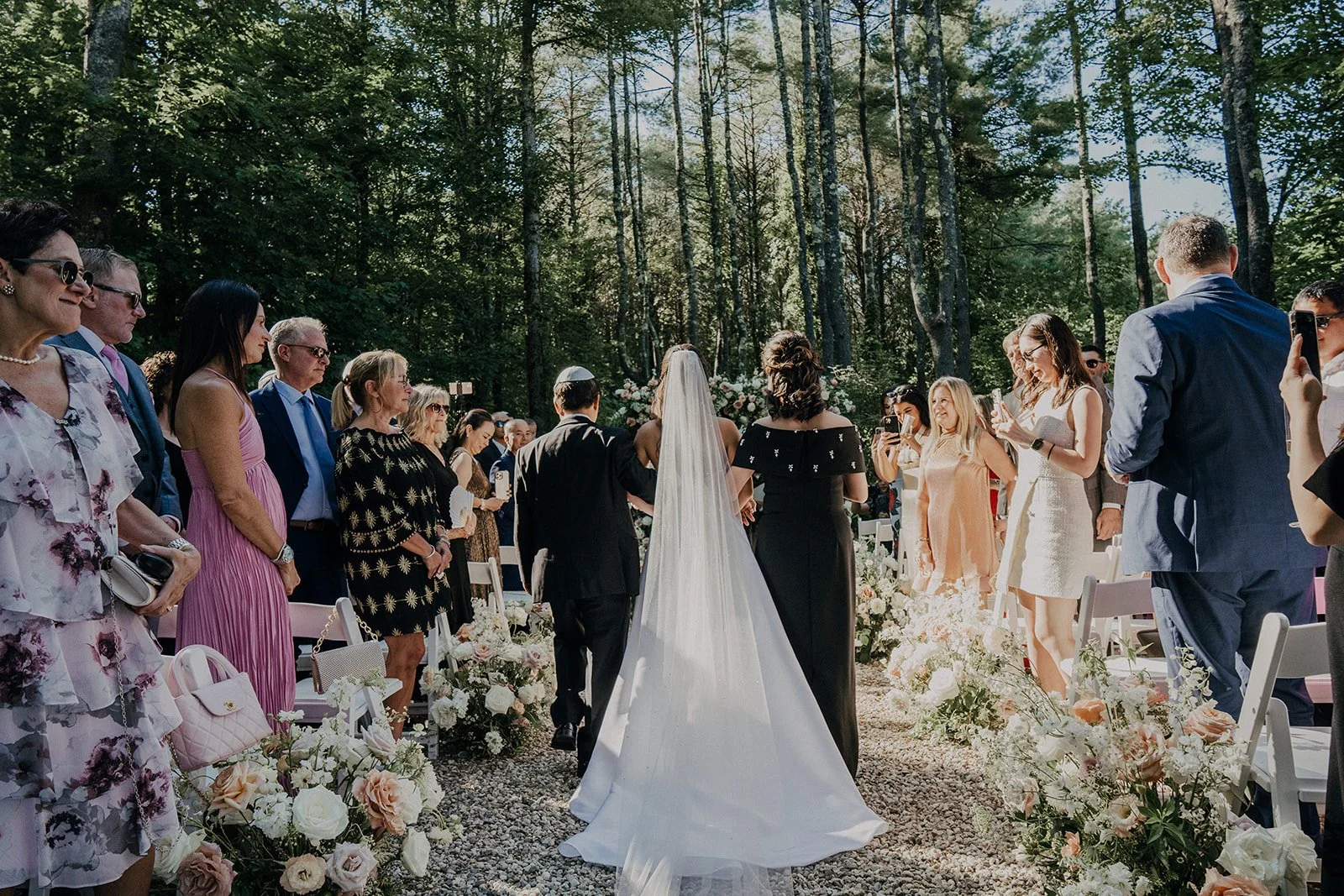 Bride and groom holding hands during their outdoor wedding ceremony, surrounded by seated guests under a forest canopy.
