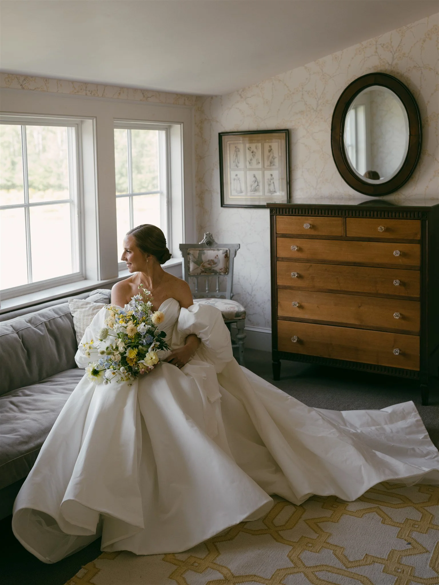 A bride in a white wedding dress sitting on a sofa and looking out the window, holding a bouquet of flowers, in a well-lit room with vintage decor.