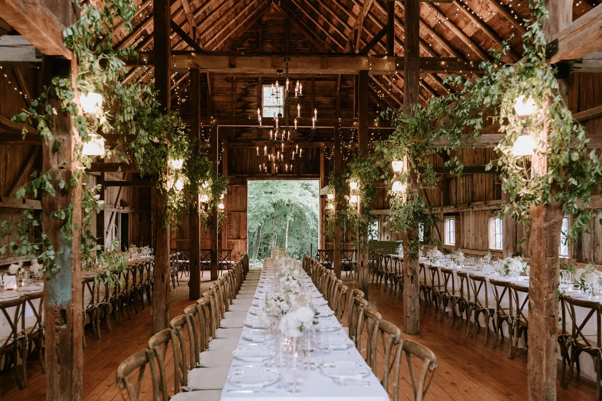 Rustic barn interior decorated for a wedding or special event with long dining tables, white floral centerpieces, greenery, and hanging string lights and chandeliers.