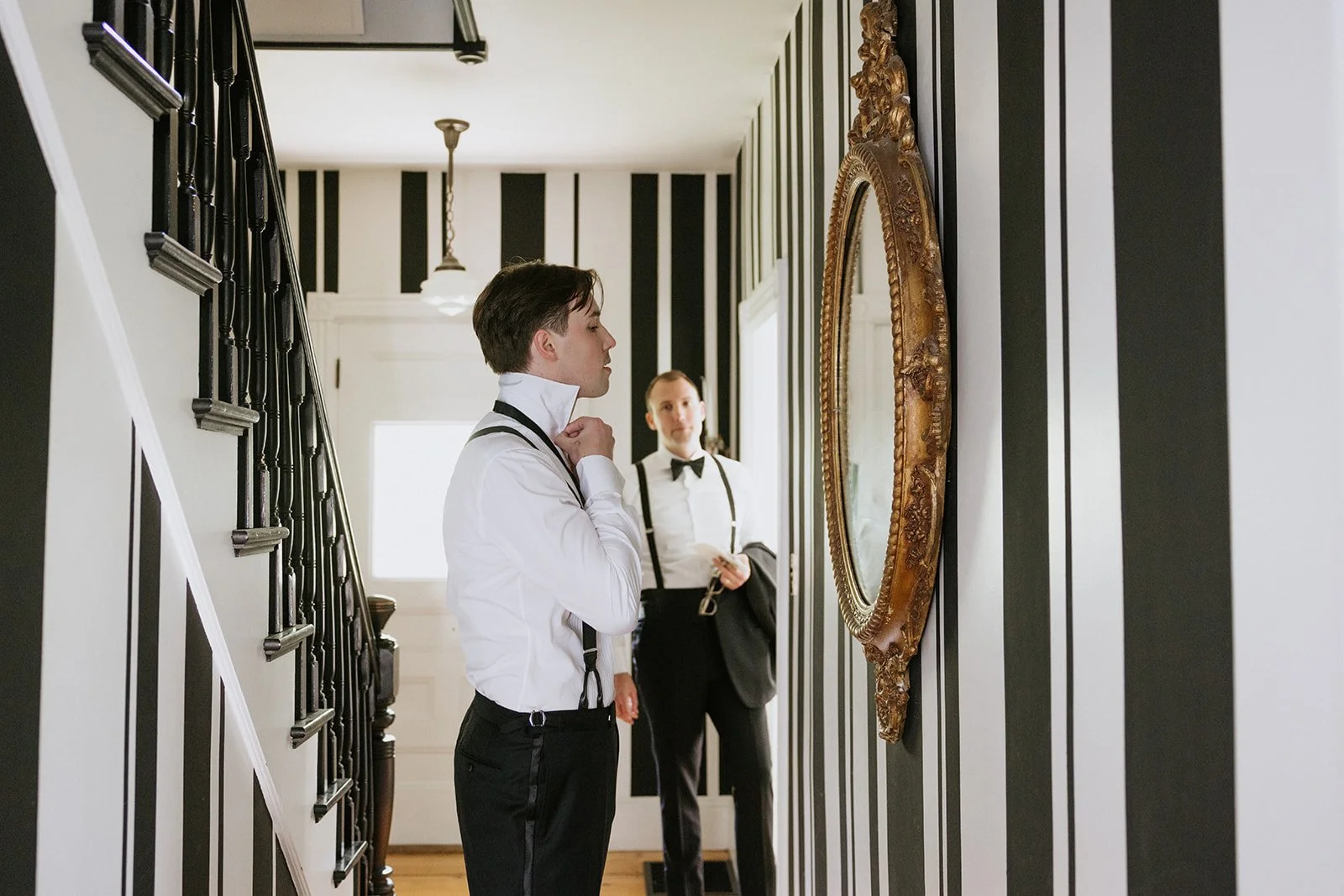 A man in formal attire, adjusting his collar, looking at a mirror in a hallway with black and white striped walls, while a woman in formal attire stands in the background.
