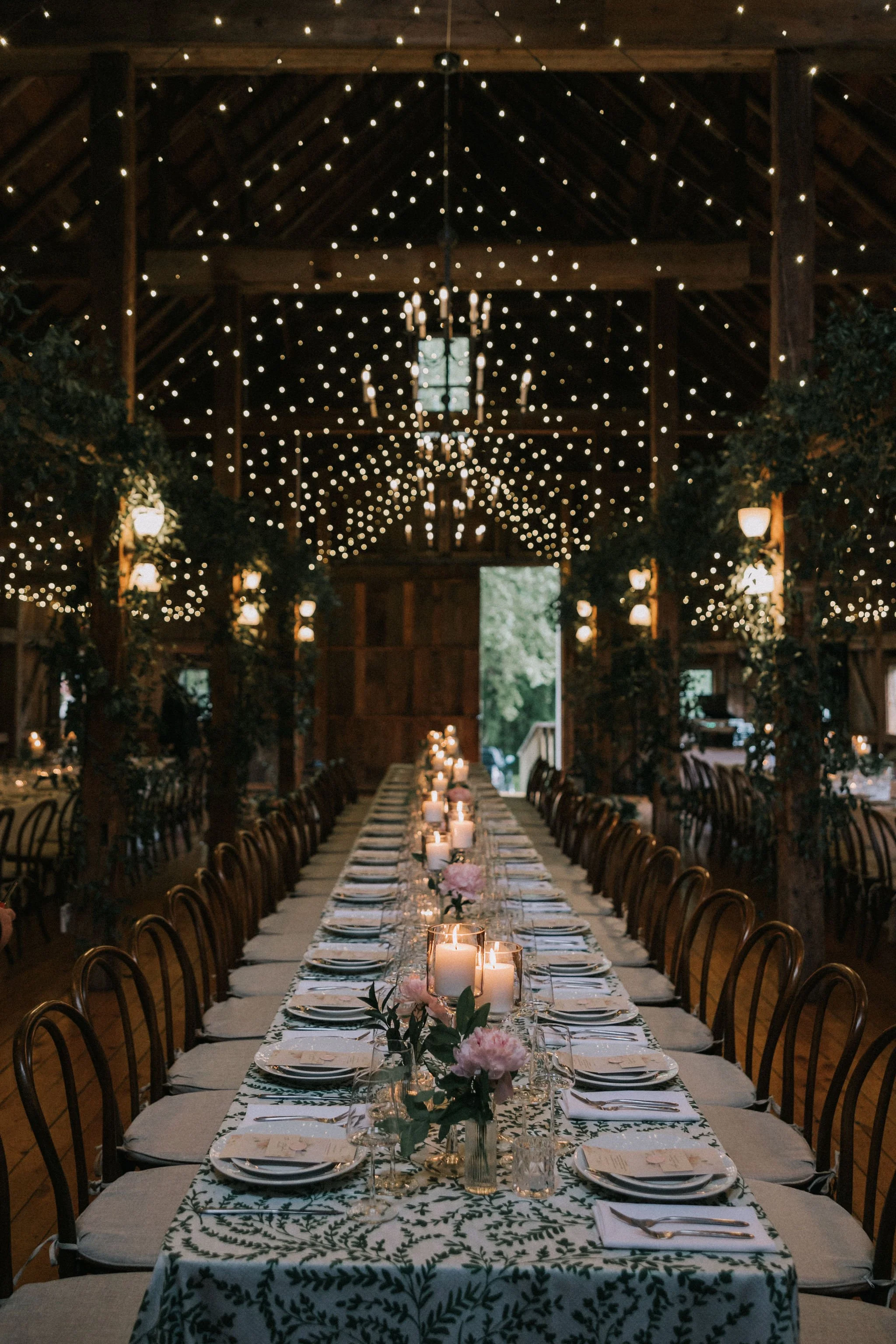 Long banquet table decorated with candles, flowers, and tableware in a rustic barn with string lights overhead for an event.