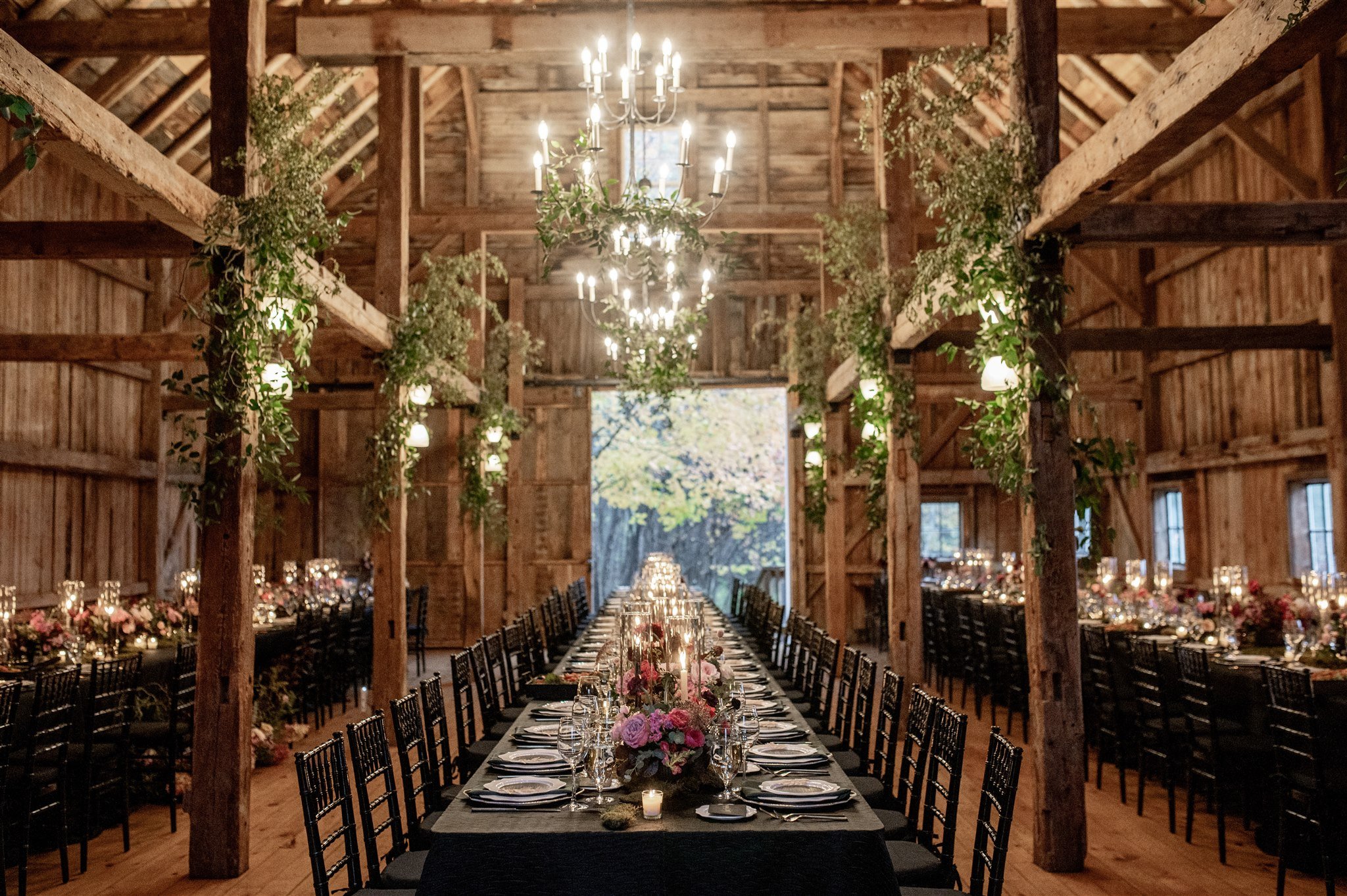 Long banquet table decorated with flowers and candles in a rustic wooden barn with chandeliers and hanging greenery