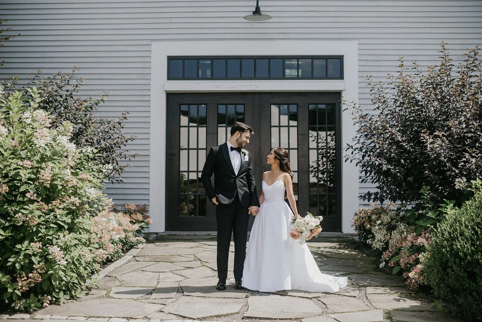 A bride and groom standing hand in hand outside a building with black double doors, surrounded by flowers and bushes.