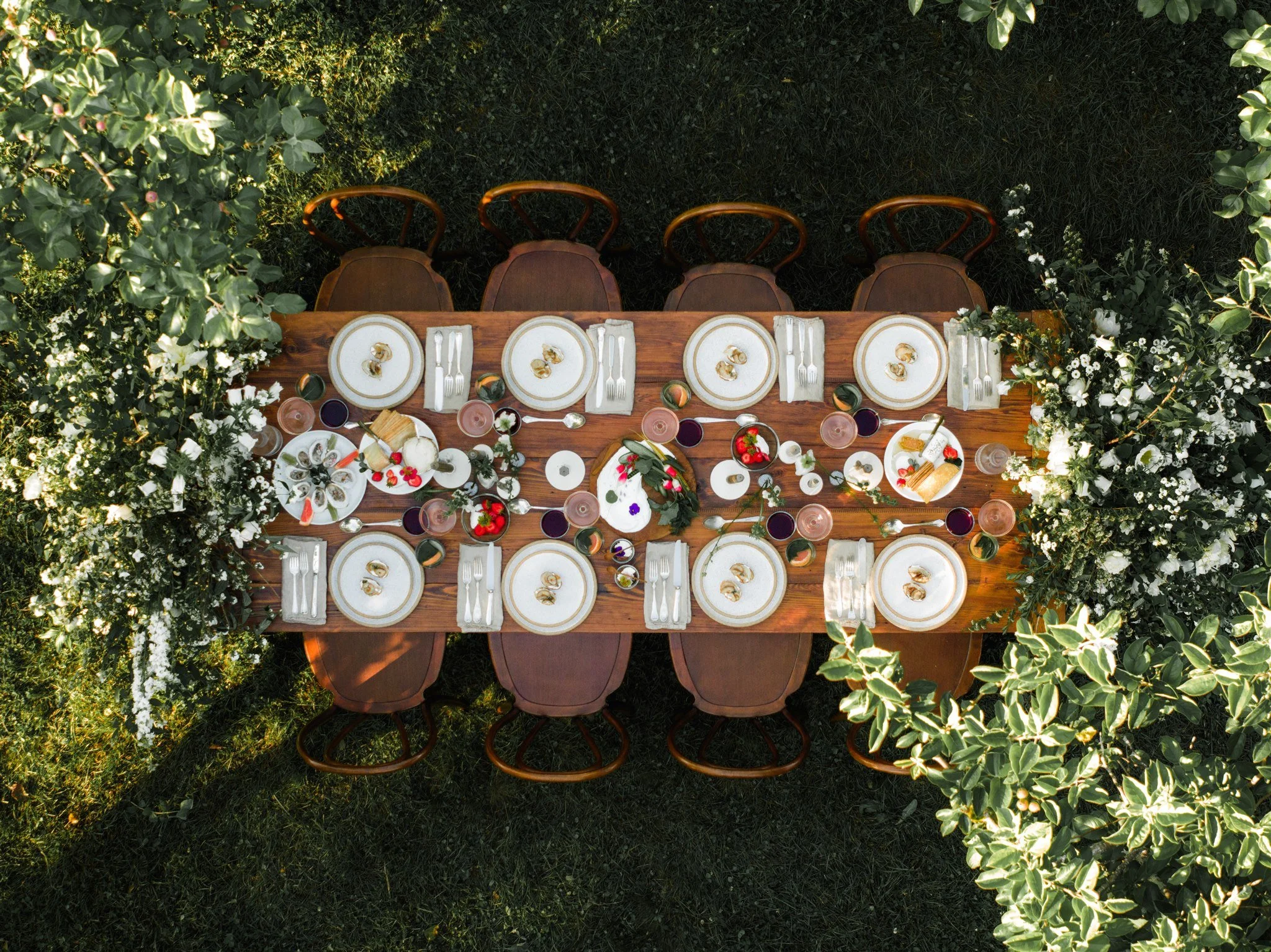 A top-down view of a long wooden outdoor dining table set with white plates, silverware, glassware, and small food items, surrounded by lush greenery and white flowers.