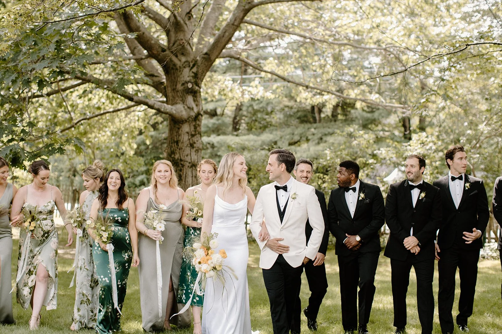 A wedding party walking outdoors under a large tree, with women in dresses and men in tuxedos, smiling and enjoying the moment.