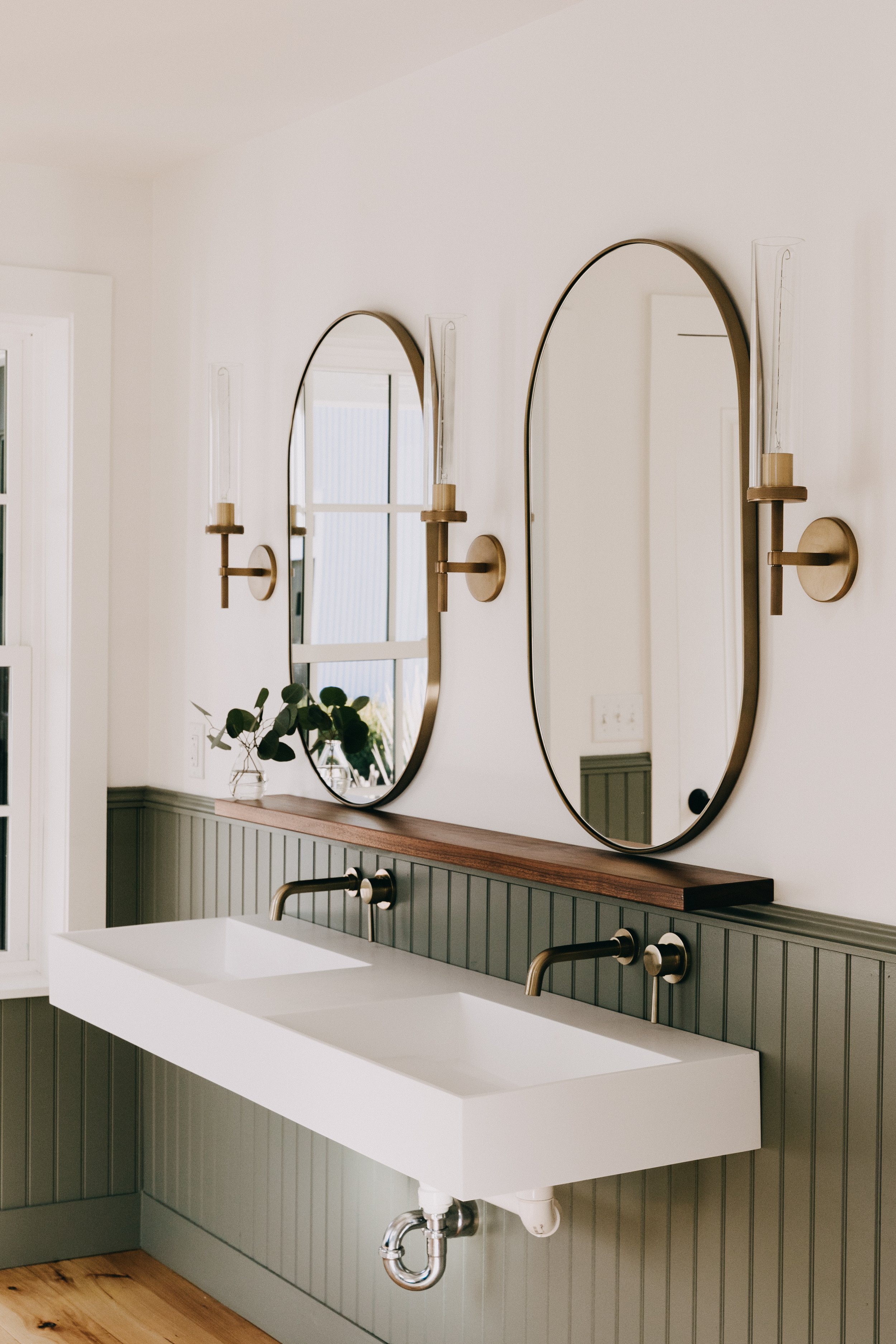Modern bathroom with dual rectangular white sinks, brass fixtures, oval mirrors, green beadboard wall, and wall-mounted candle sconces.