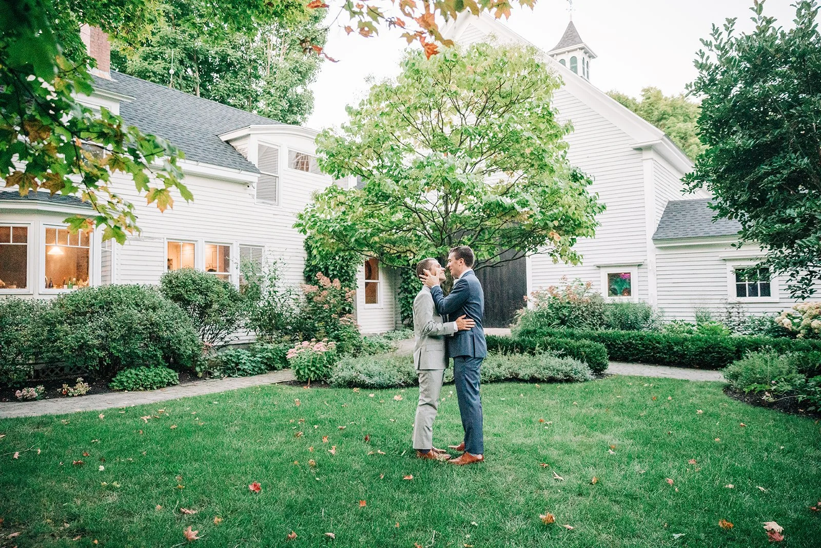 A couple in suits standing in a green backyard, embracing and smiling at each other, with a white house and trees in the background.