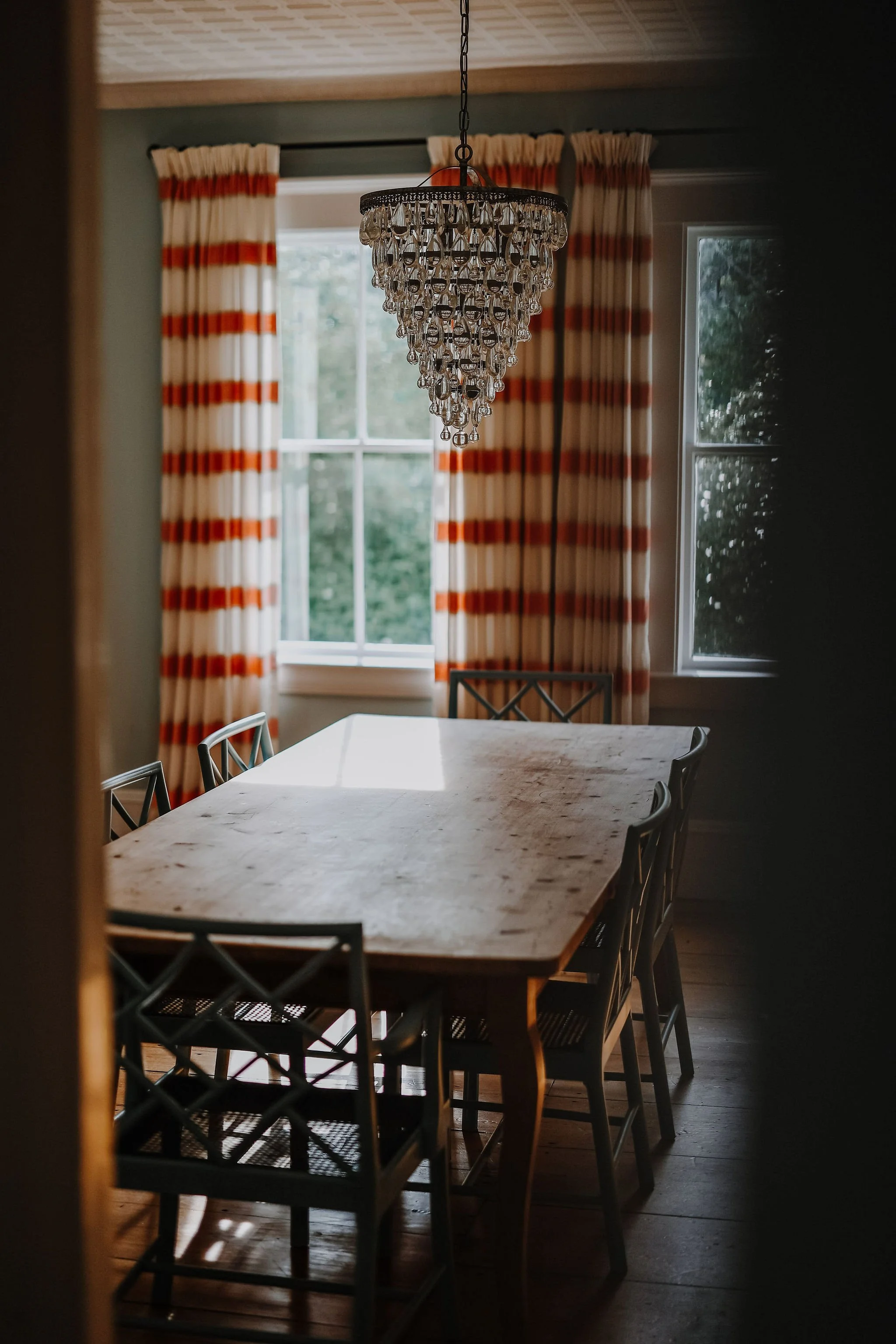 A wooden dining table with eight chairs, a crystal chandelier hanging above, and windows with cream curtains with orange and red stripes in the background.