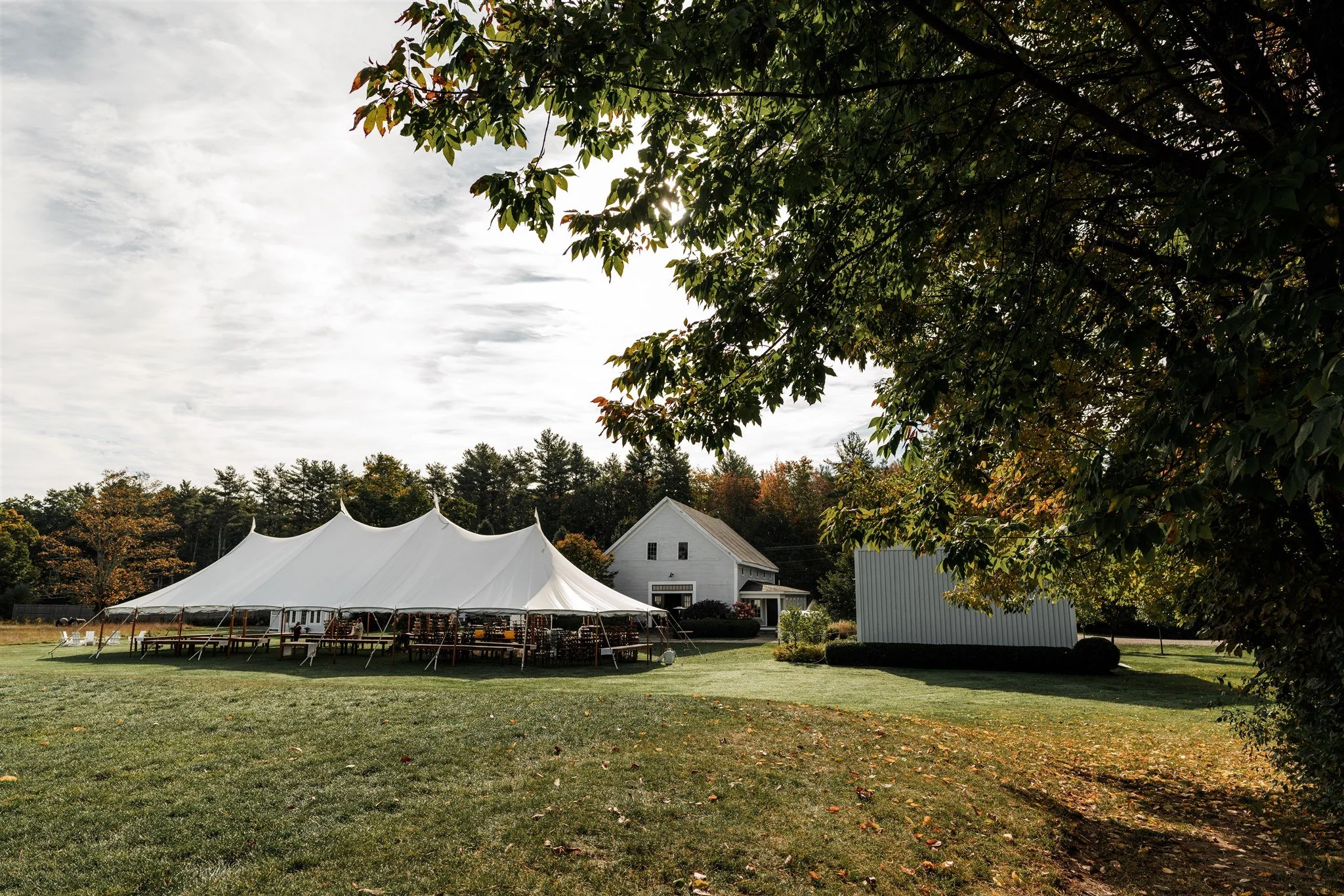 A large white event tent set up on a grassy field with tables underneath, near a white house with a gray roof, surrounded by trees with fall foliage. Overcast sky.