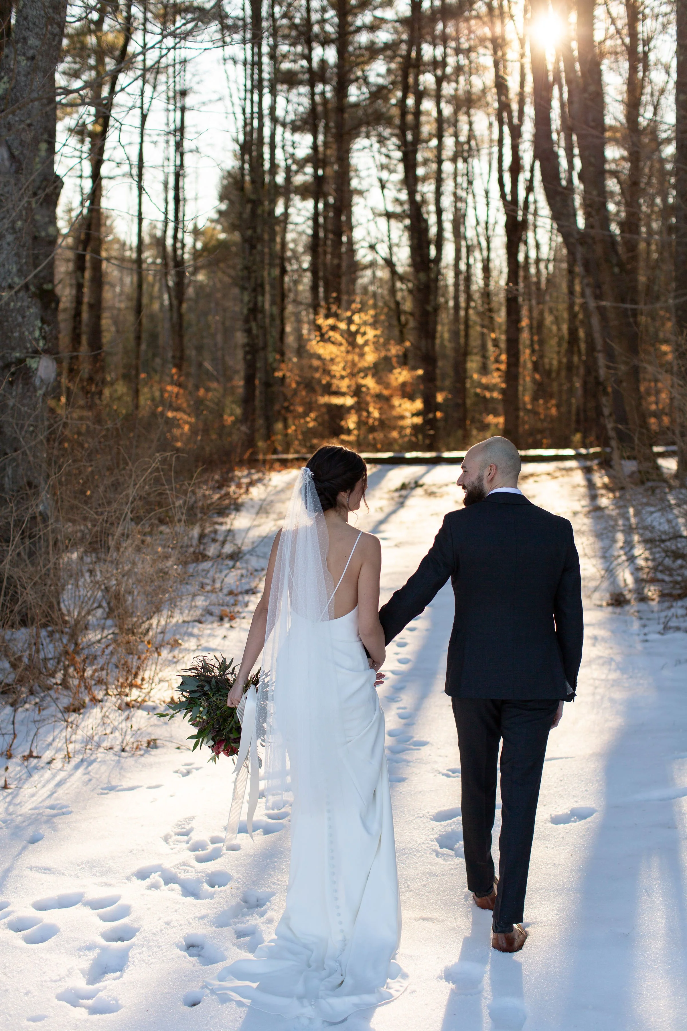 A bride and groom walking hand in hand through a snowy forest during sunset, with the bride holding a bouquet and wearing a white wedding dress with a veil, and the groom in a dark suit.