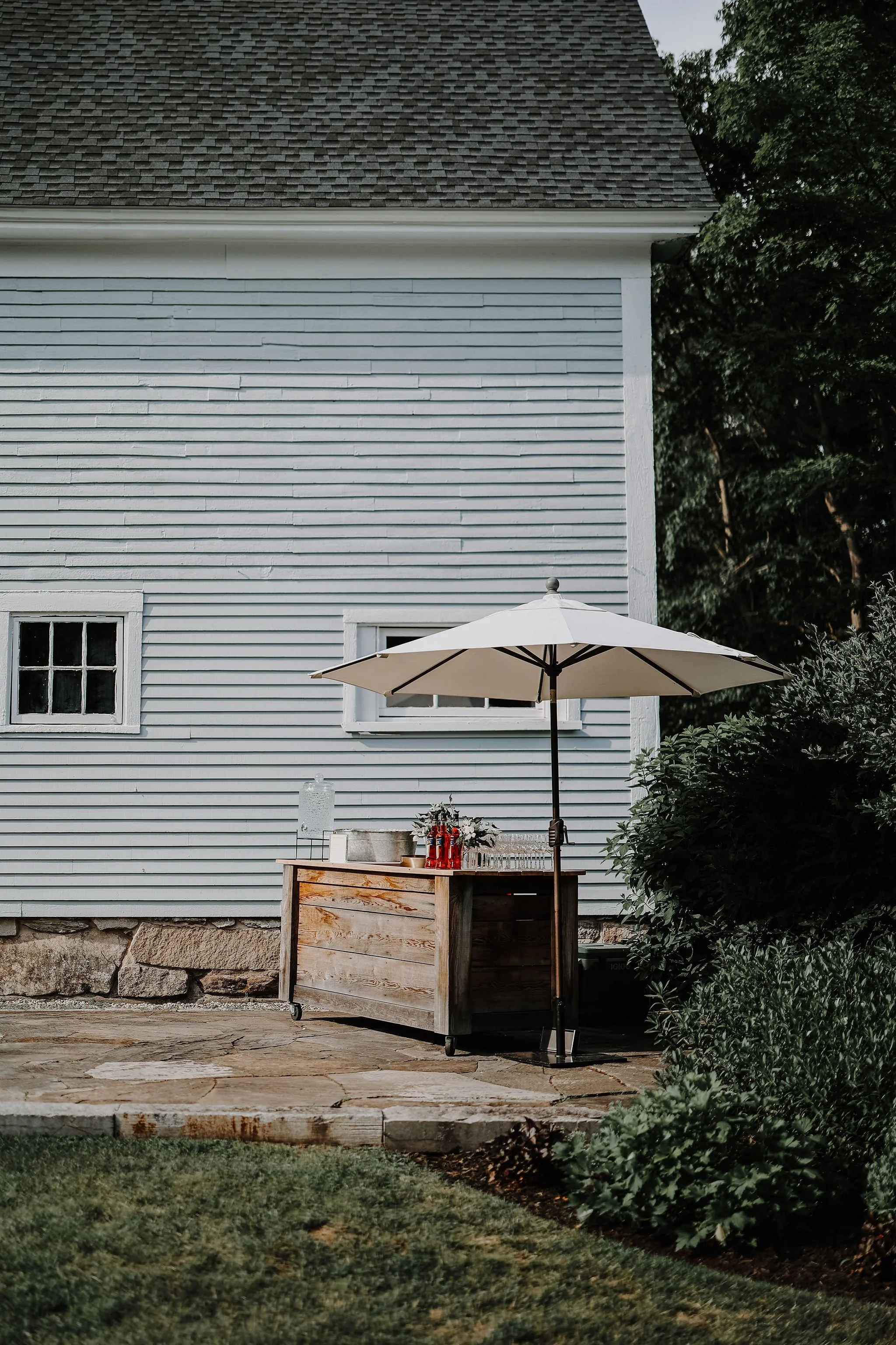 An outdoor setup next to a house with light blue siding and a dark shingled roof. There is a large beige umbrella over a wooden bar with drinks and glasses on it. Green bushes and trees surround the area.