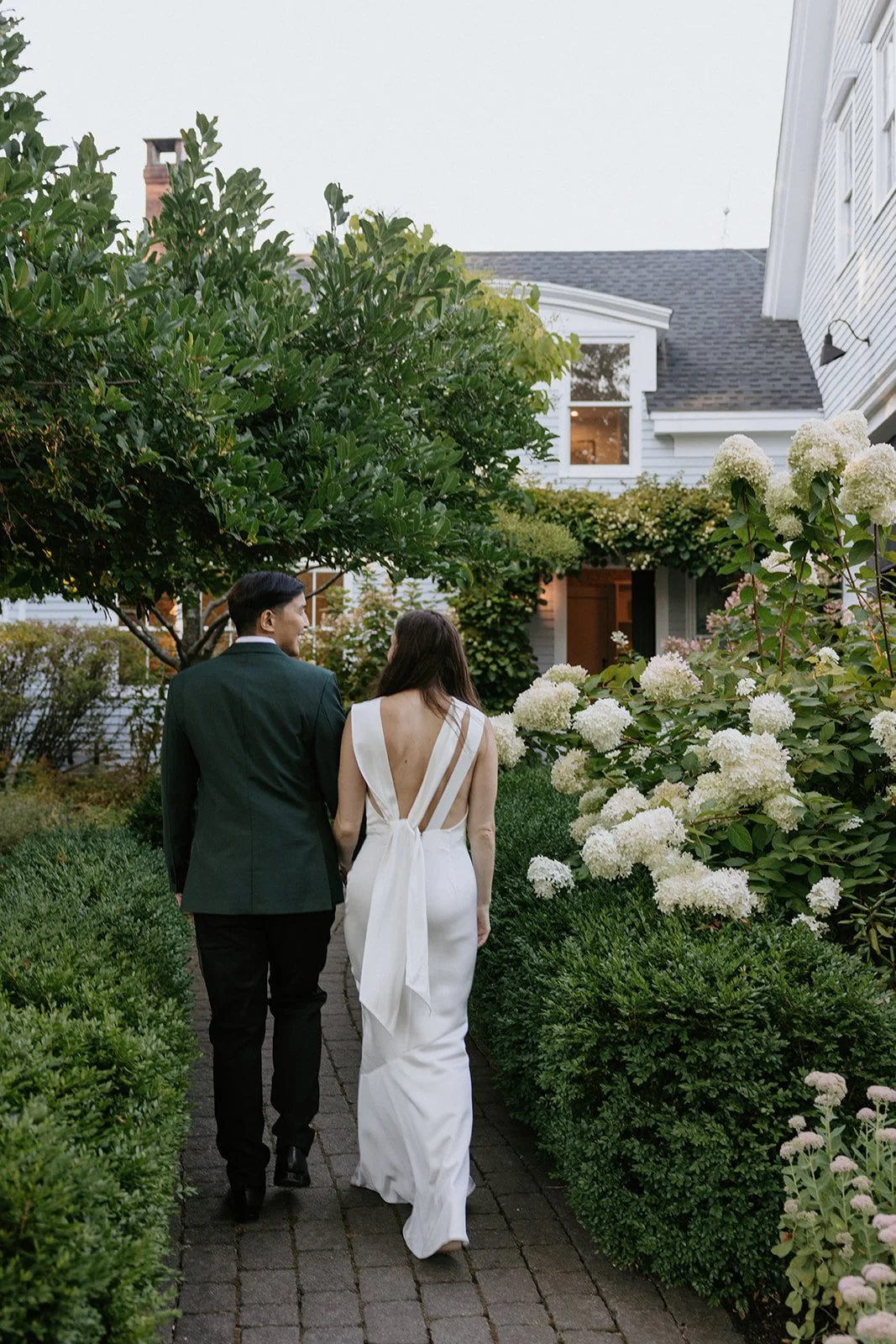 A couple, dressed in formal attire, walking on a garden path with lush greenery and white flowering bushes in the evening.