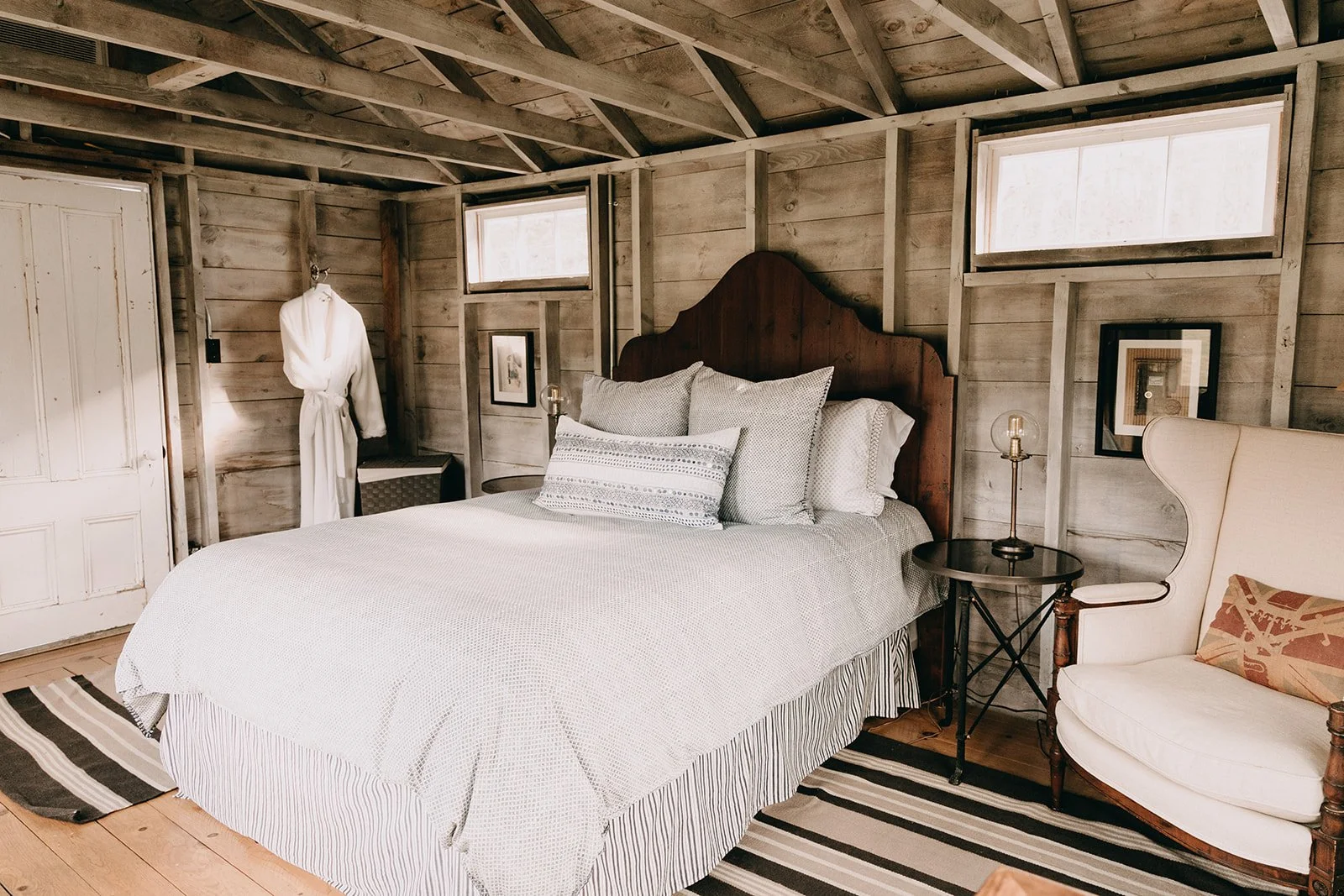 Rustic bedroom with a wooden-paneled wall, a bed with multiple pillows, a nightstand with a lamp, a cozy armchair, and two small horizontal windows.