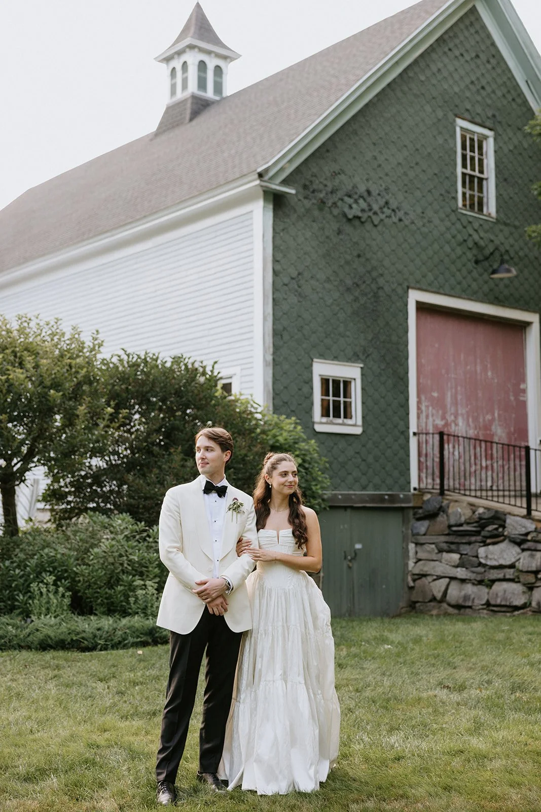 A bride and groom standing outdoors on a grassy area, with a large green and white barn in the background. The groom is dressed in a white tuxedo jacket with a black bow tie, and the bride is wearing a strapless white wedding gown. They are holding o