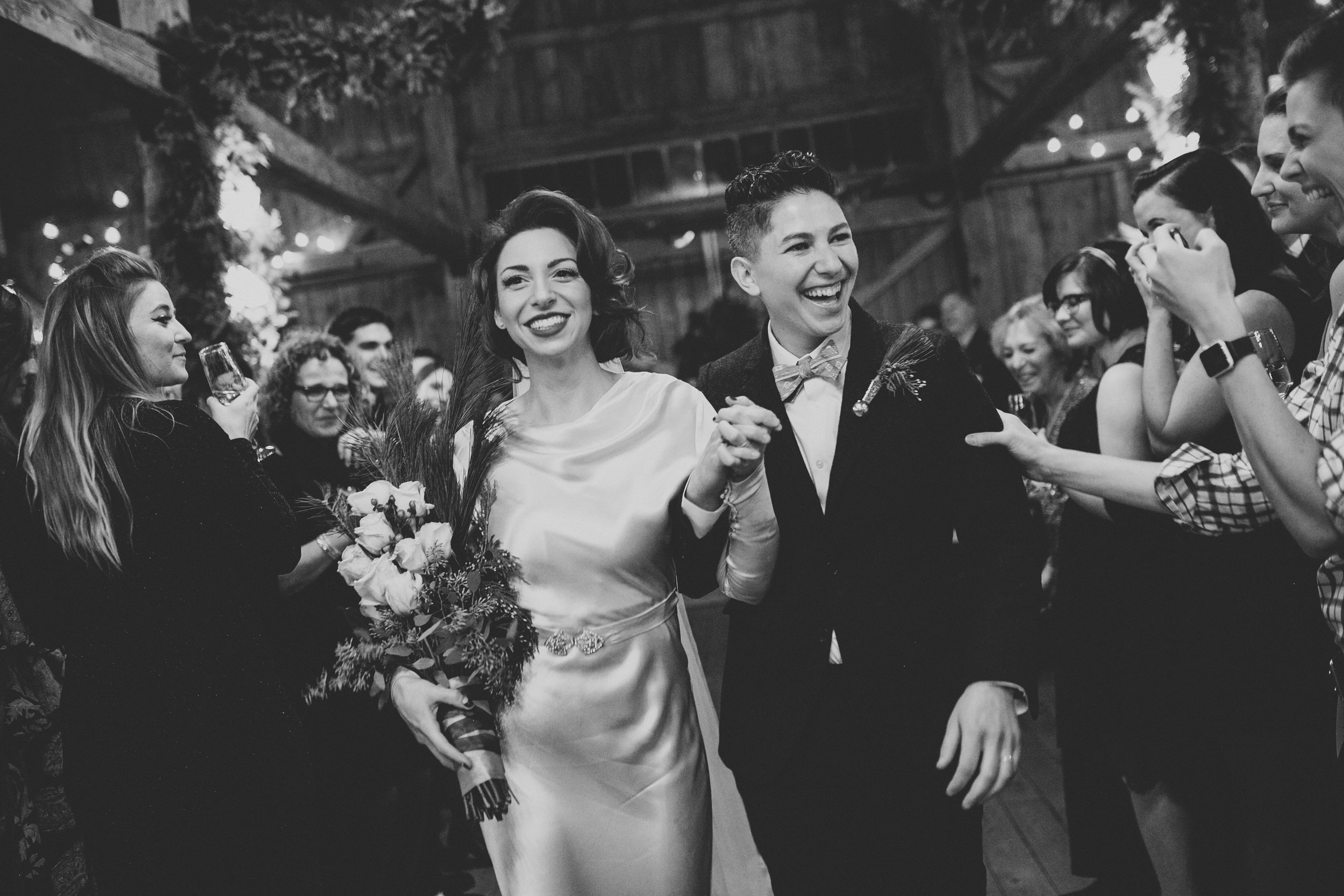A bride and groom smiling and holding hands during their wedding reception, surrounded by guests celebrating in a rustic indoor venue.