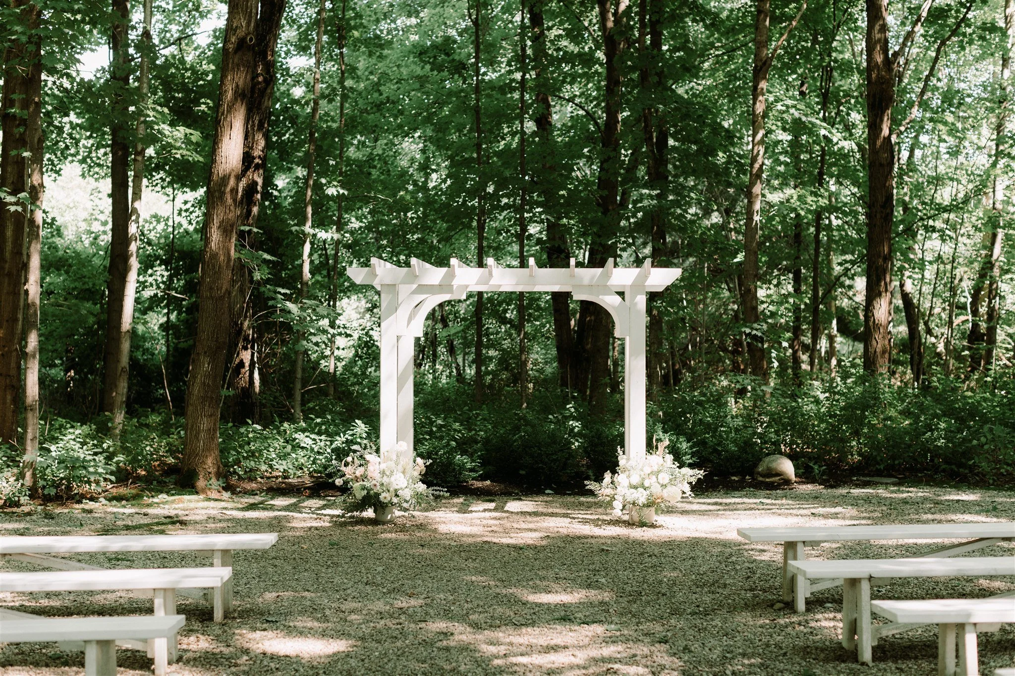 A white wedding arch adorned with floral arrangements is set outdoors among green trees, with white benches on either side.