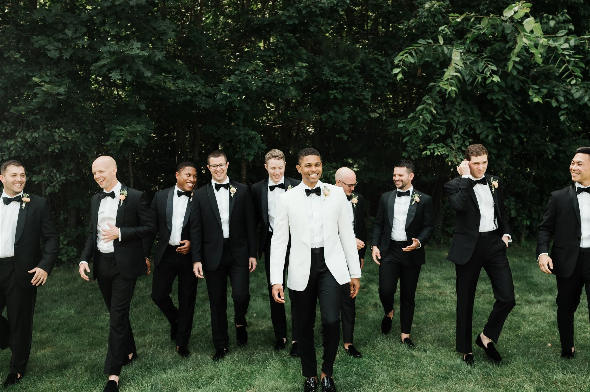 A group of men in tuxedos walking on grass. The man in the white tuxedo is smiling, surrounded by men wearing black tuxedos with bow ties, with some laughing and others smiling, against a background of dense trees.