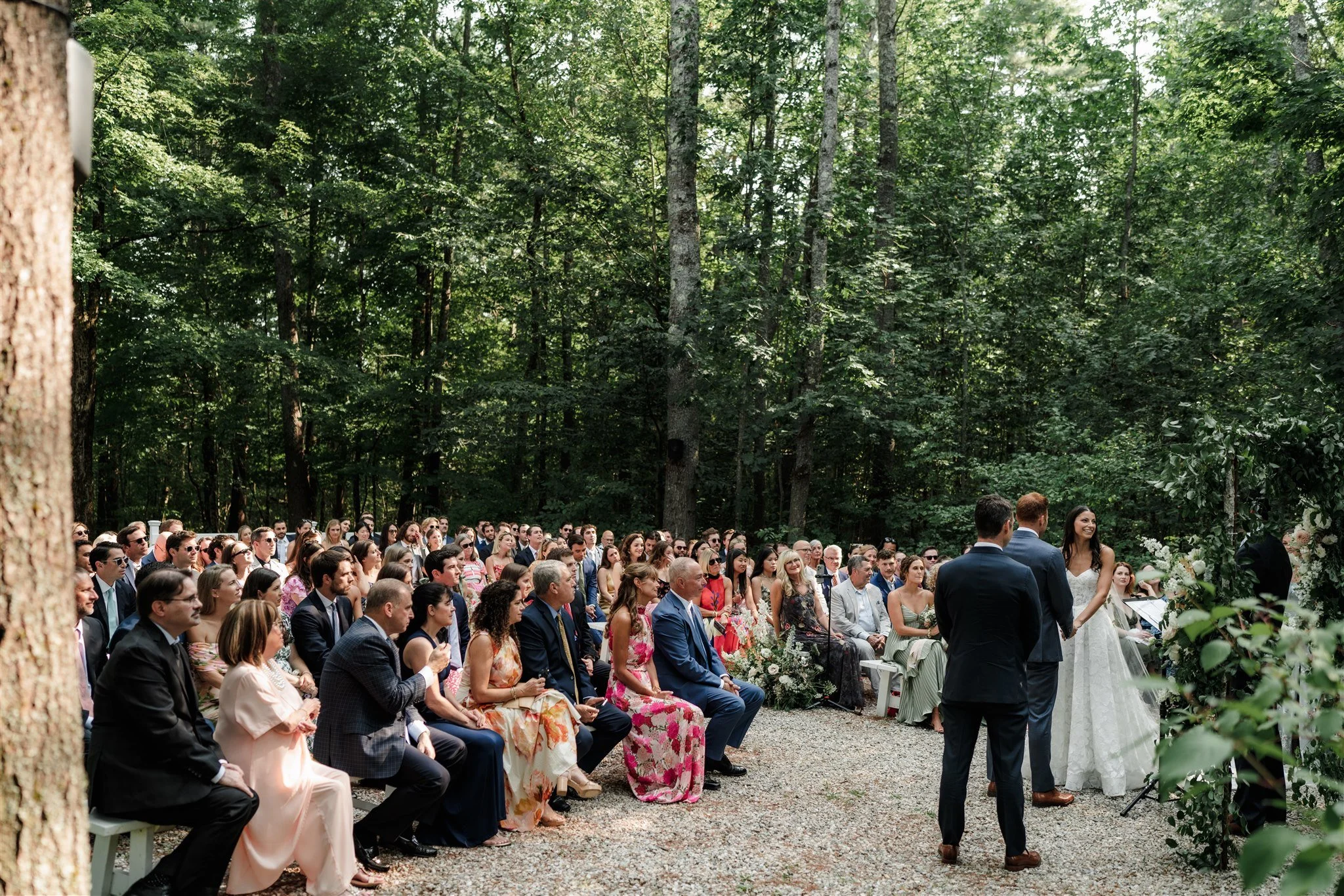 A wedding ceremony taking place outdoors in a wooded area with a large group of guests seated and watching the bride and groom exchange vows.