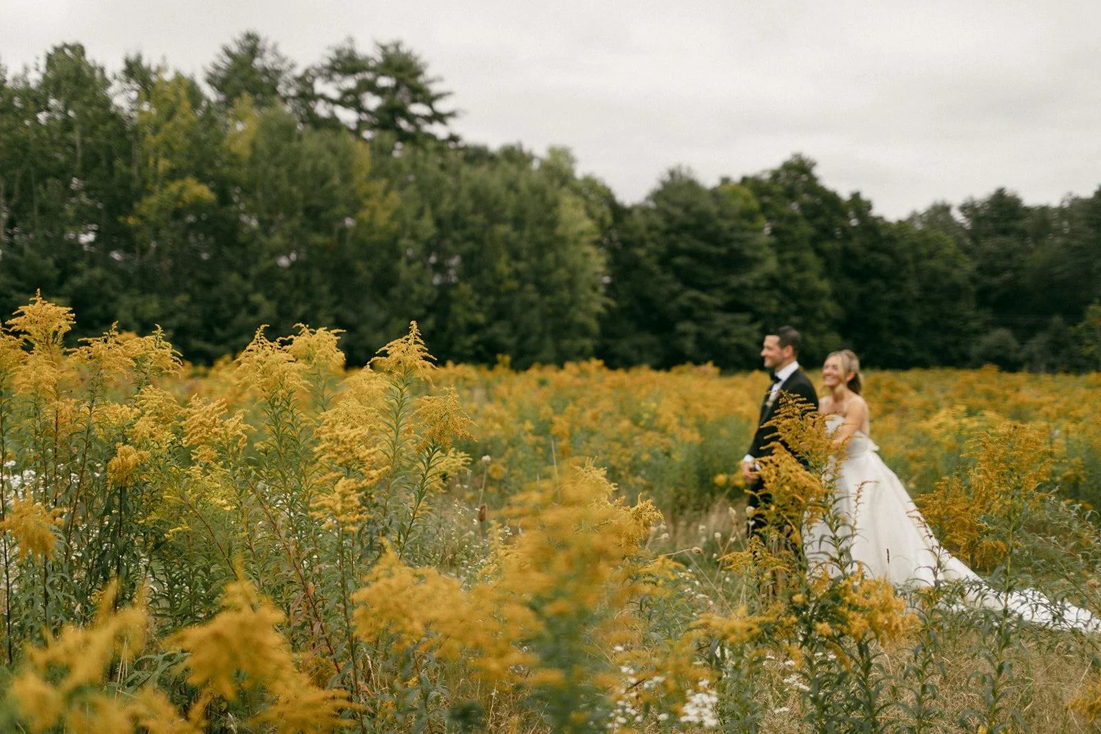 A bride and groom standing in a yellow wildflower field with trees in the background, smiling.