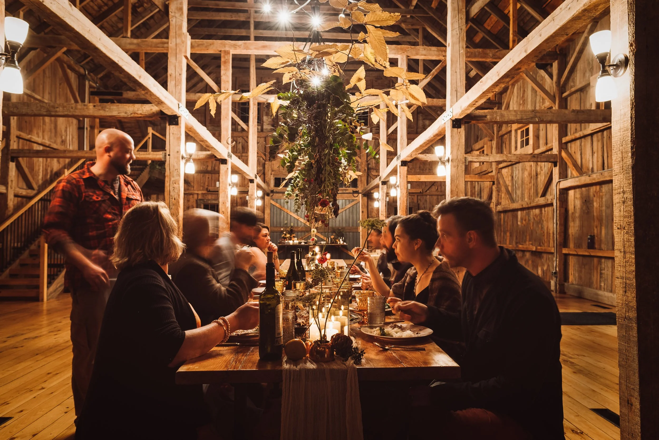 A group of people gathered around a dinner table in a rustic barn, decorated with hanging foliage, candles, and bottles, with warm lighting and wooden beams overhead.
