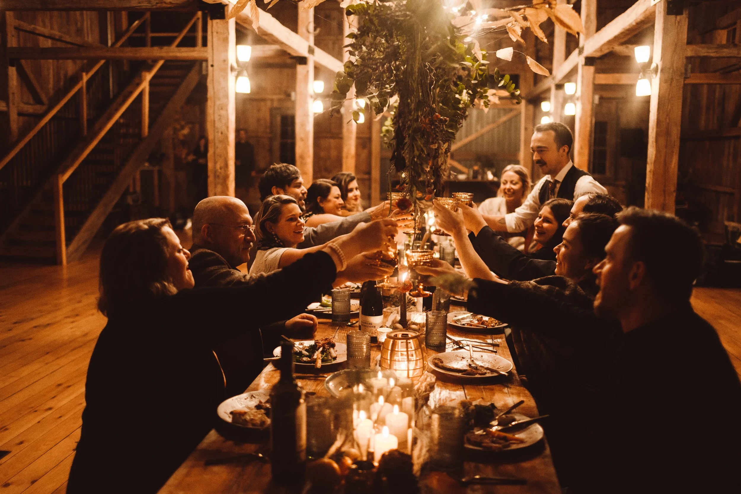 A group of people gathered around a long wooden table inside a rustic barn, celebrating with drinks and toasts under warm hanging lights and candles.