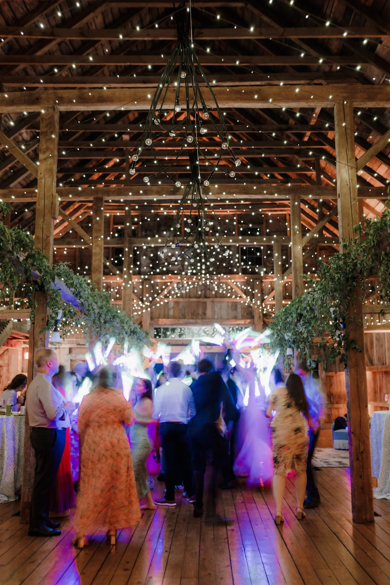 People dancing and socializing in a rustic barn decorated with string lights and greenery, with a DJ mixing music and colorful lighting effects.