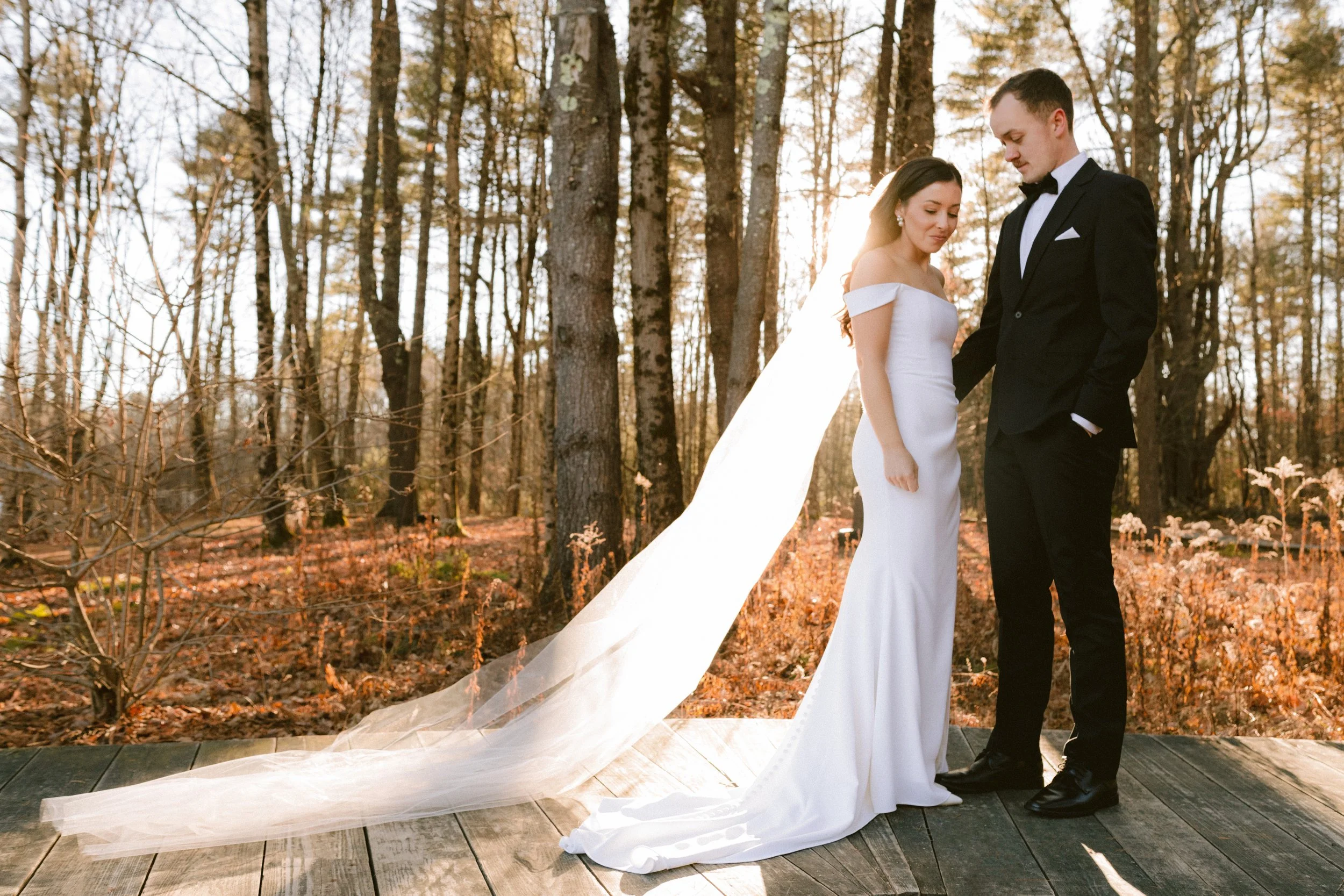 Bride and groom standing close together on a wooden platform in a forest with autumn leaves, the bride in a white off-shoulder wedding gown with a long train, and the groom in a black tuxedo with a bow tie, sunlight shining through the trees.