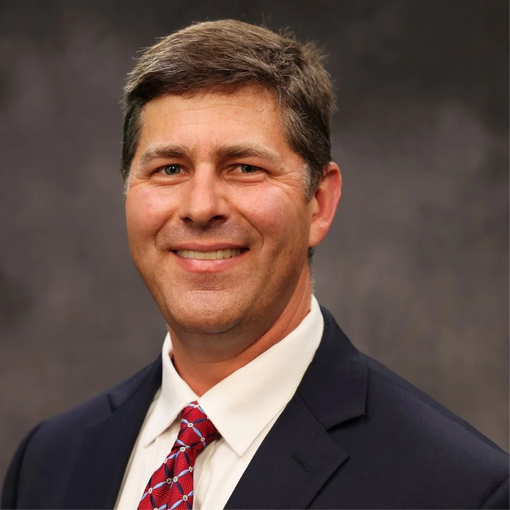 Portrait of a man wearing a dark suit, white shirt, and red patterned tie, smiling against a dark background.