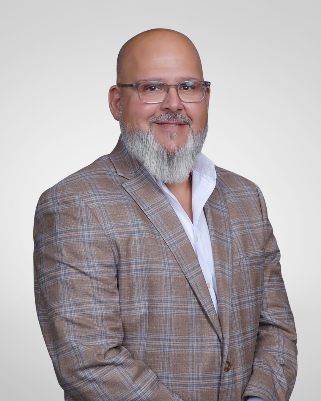 A man with glasses and a gray beard wearing a brown checked blazer and white shirt, standing against a plain gray background.
