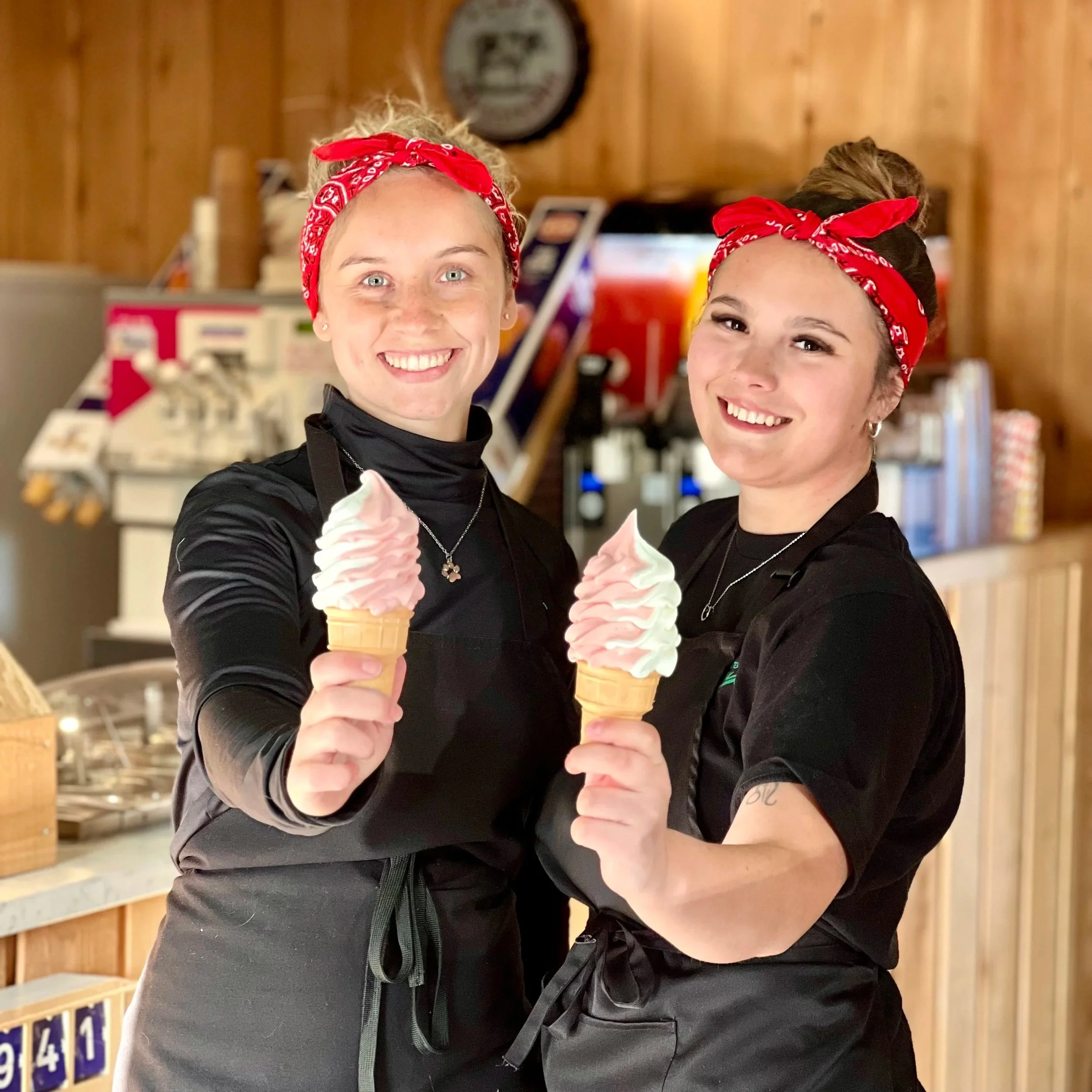 Deux femmes souriantes portant des bandanas rouges et des tabliers noirs, tenant des cornets de glace à la crème glacée à la vanille et à la fraise dans un café ou une crèmerie en bois.