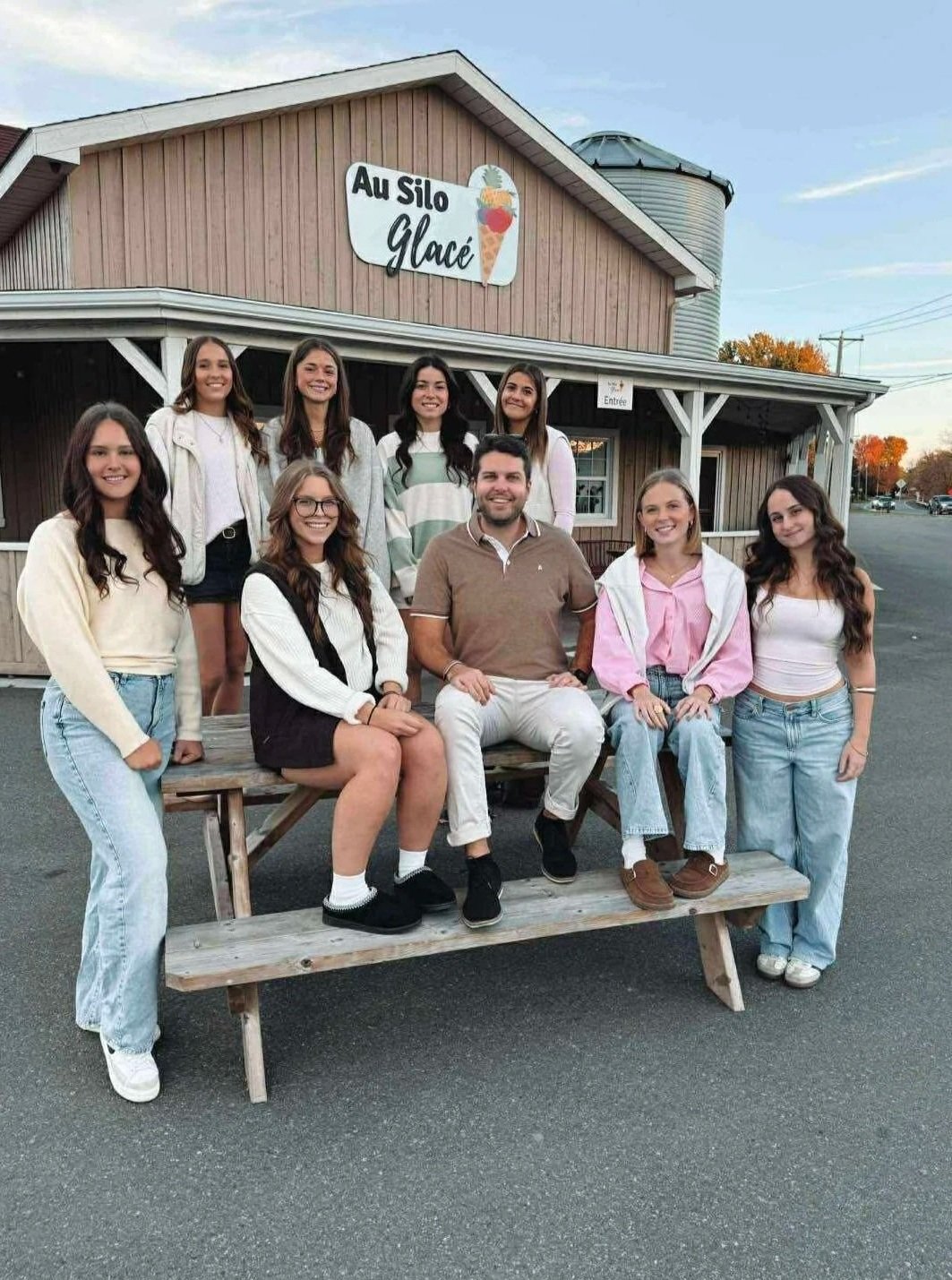 Groupe de huit jeunes femmes et un homme posant devant un bâtiment en bois avec un panneau indiquant "Au Silo Glacé", l'automne en arrière-plan.
