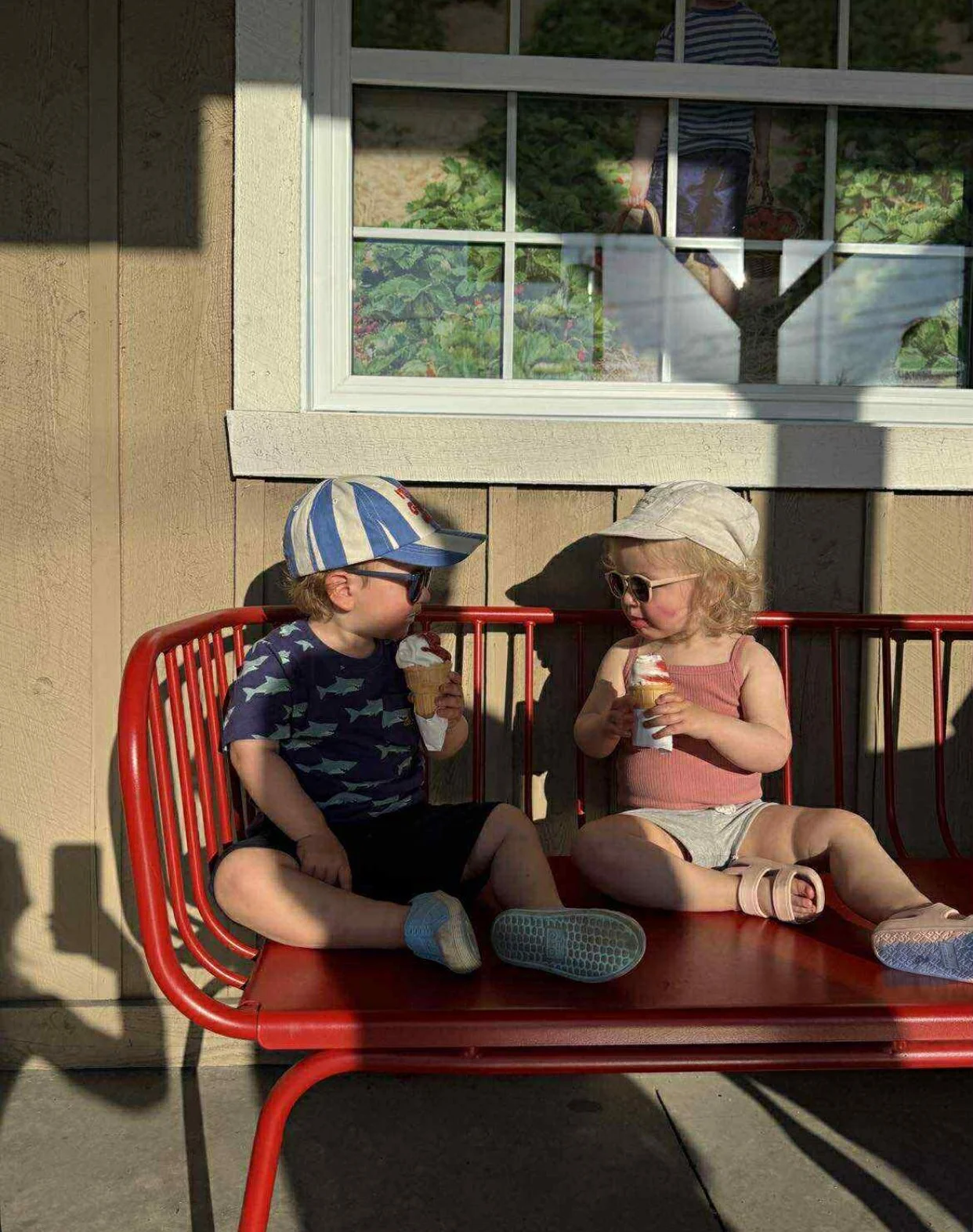 Deux enfants assis sur un banc rouge, en train de manger des glaces, en été, avec une maison en arrière-plan.