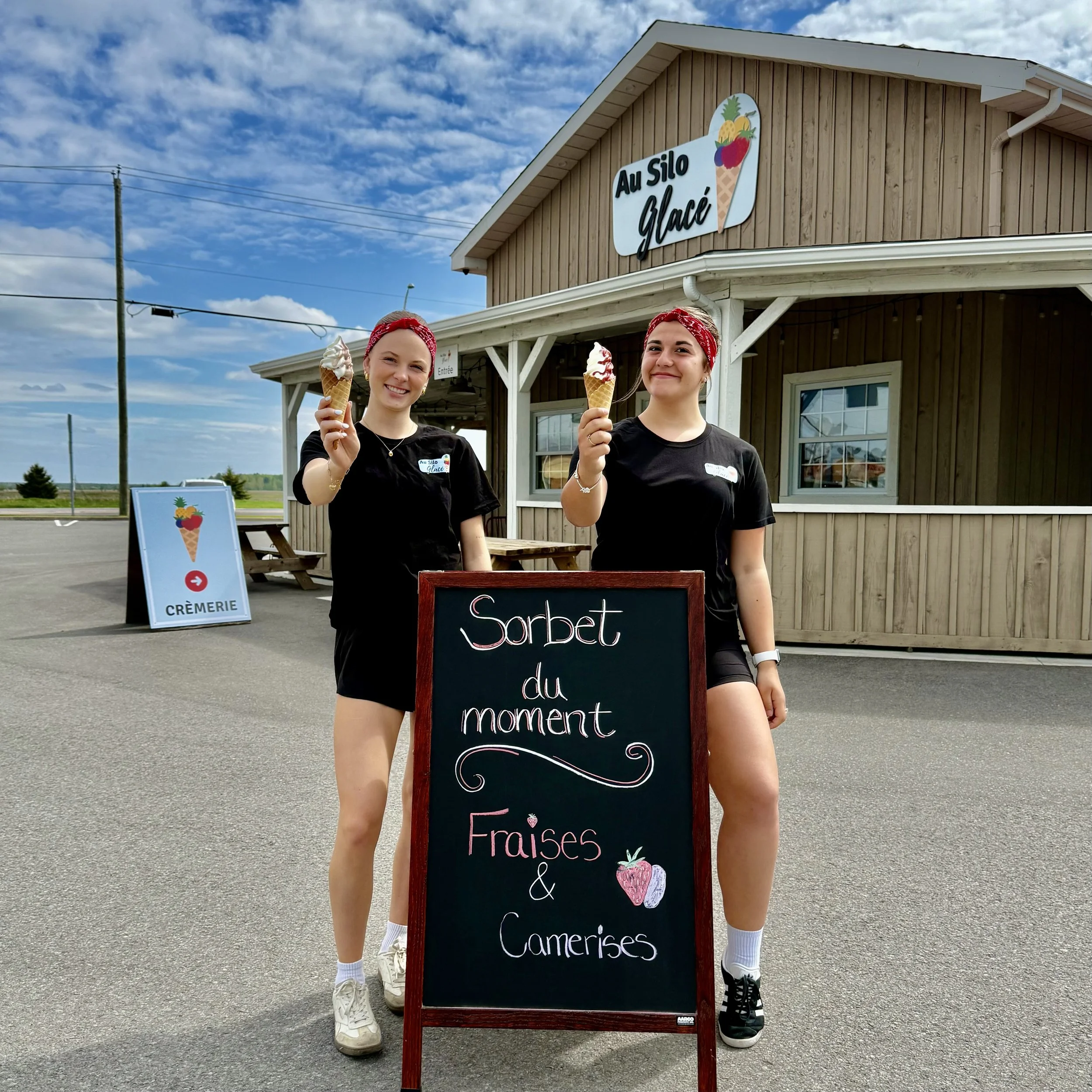 Deux jeunes femmes posent devant un stand de crème glacée, tenant chacune une corne de glace, avec un tableau noir indiquant « Sorbet du moment Fraises & Camerises ». En arrière-plan, un bâtiment en bois avec un signe « Au Silo Glacé » et un panneau 