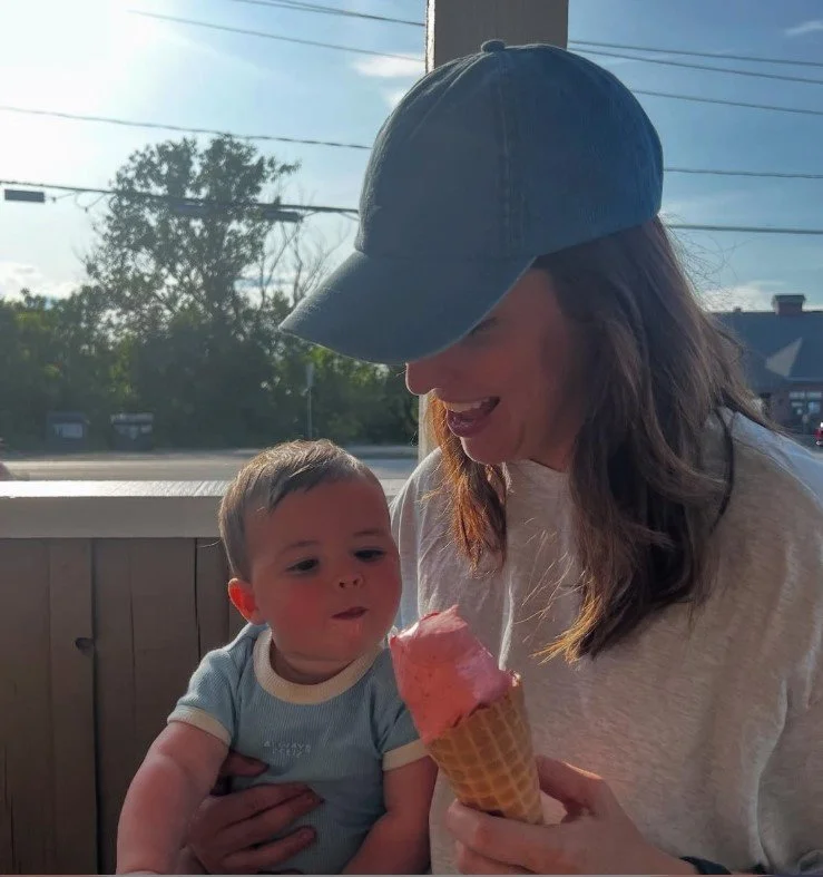 Une femme portant une casquette bleue tient un jeune enfant en main. La femme regarde un cornet de glace à la fraise, le jeune enfant regarde la glace avec curiosité, à l'intérieur d'une boutique ou d'un restaurant avec une vue sur la rue et des arbr
