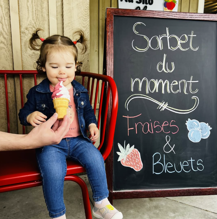 Une jeune fille assise sur un banc rouge, dégustant une glace à la fraise dans un cornet, devant un tableau noir publicitaire pour des sorbets au moment, avec des dessins de fraise, de bleuets et de petits fruits.