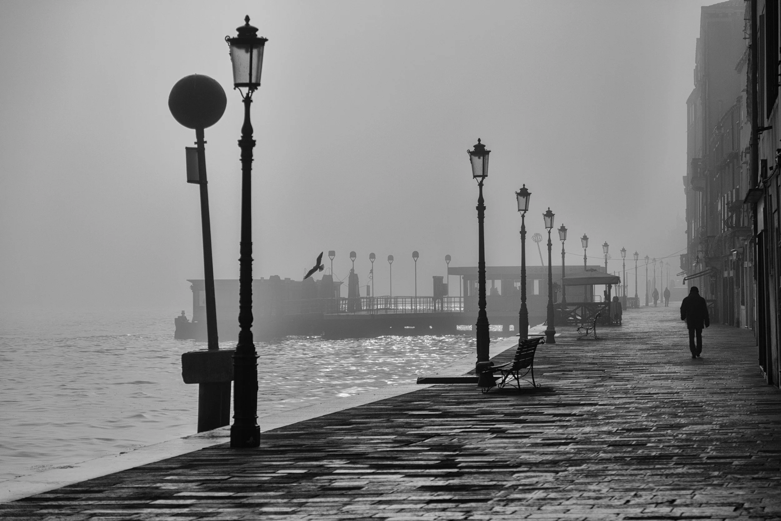 Black and white photo of a foggy waterfront promenade with vintage street lamps, benches, people walking, and a bird flying over the water.