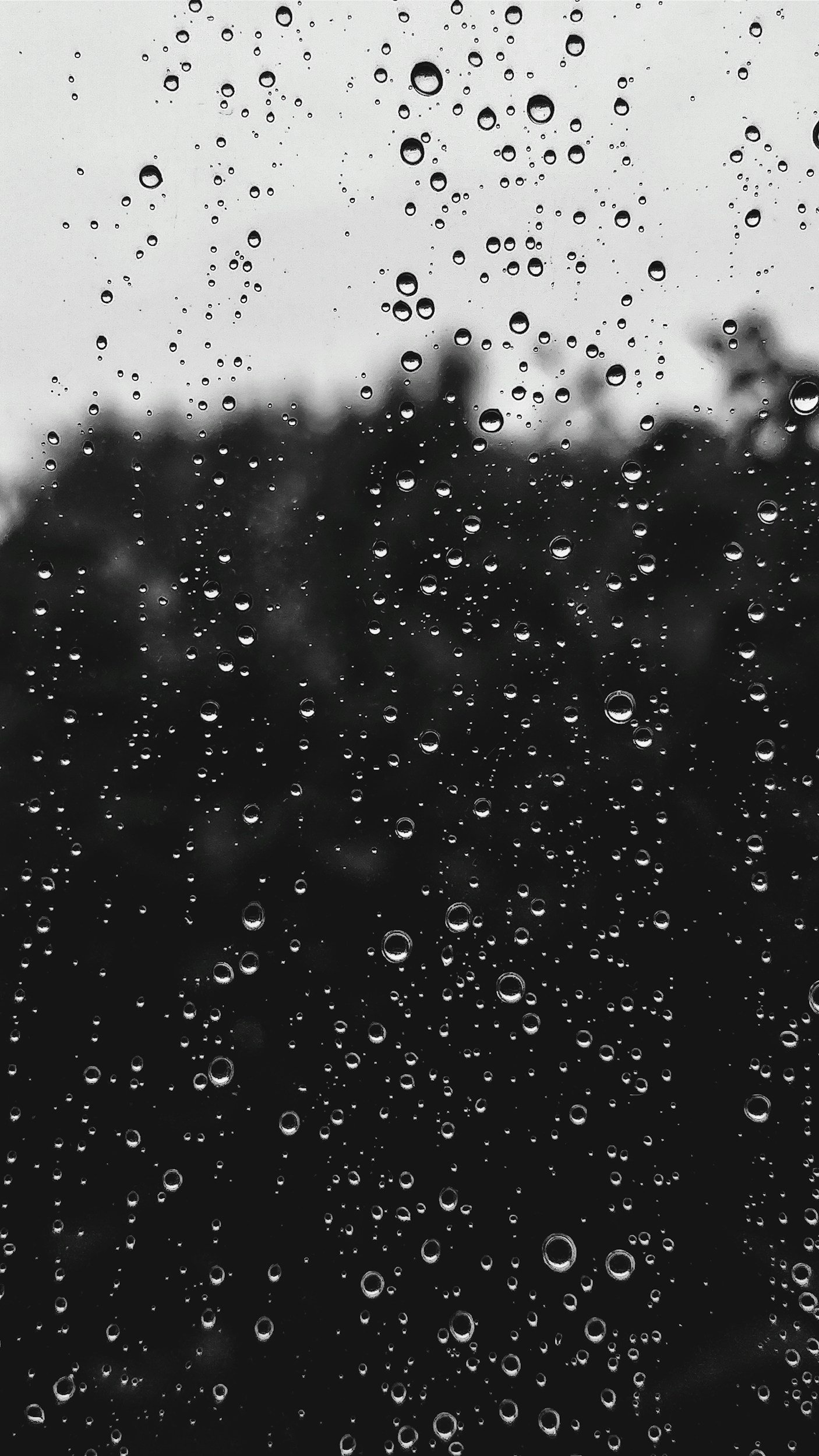 Close-up of a rain-covered window with water droplets on the glass and a blurry background of trees or bushes outside.