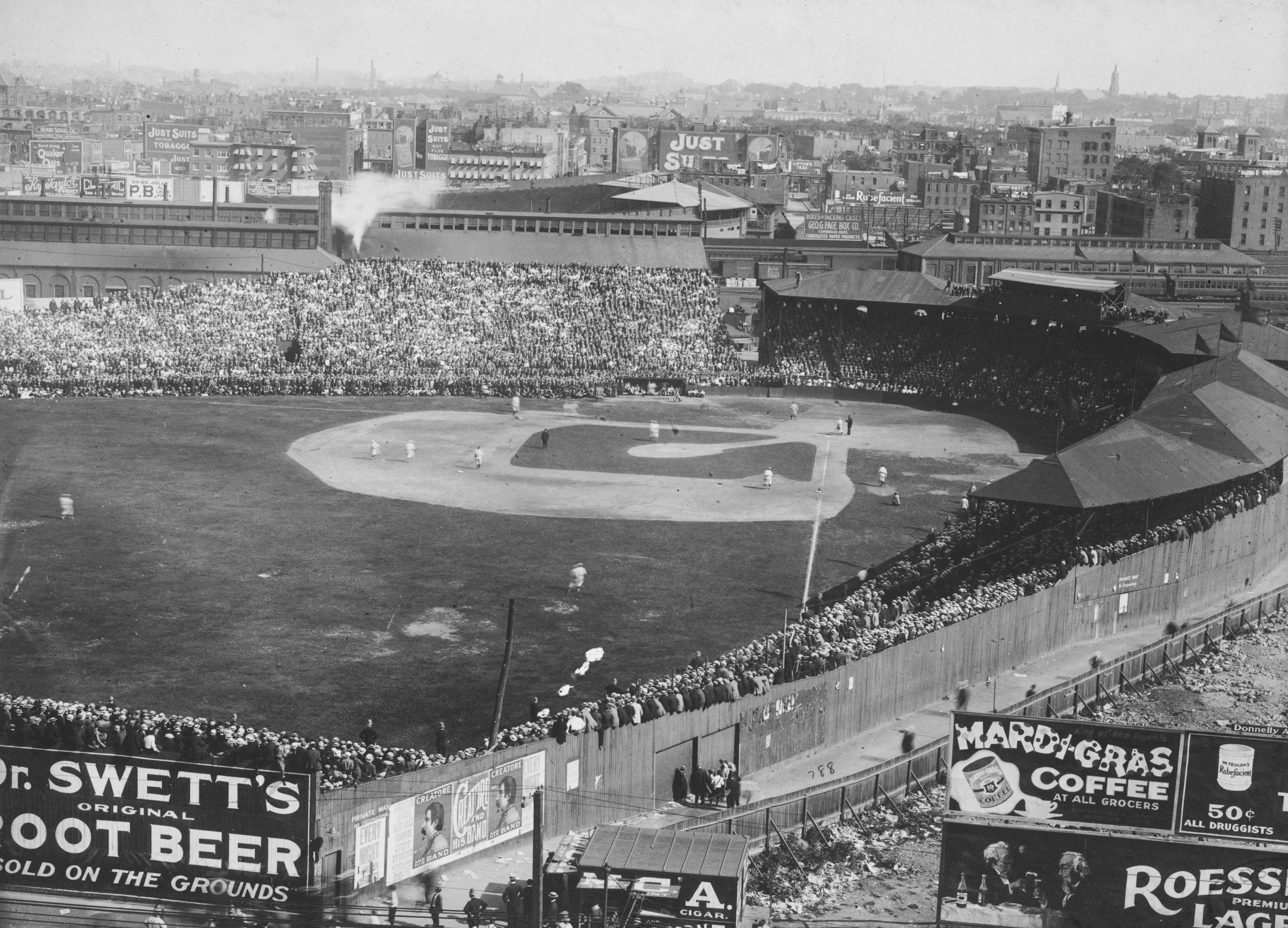 Historical black-and-white photograph of a crowded baseball stadium with players on the field and spectators in the stands. The city skyline with multiple billboards and advertisements is visible in the background.