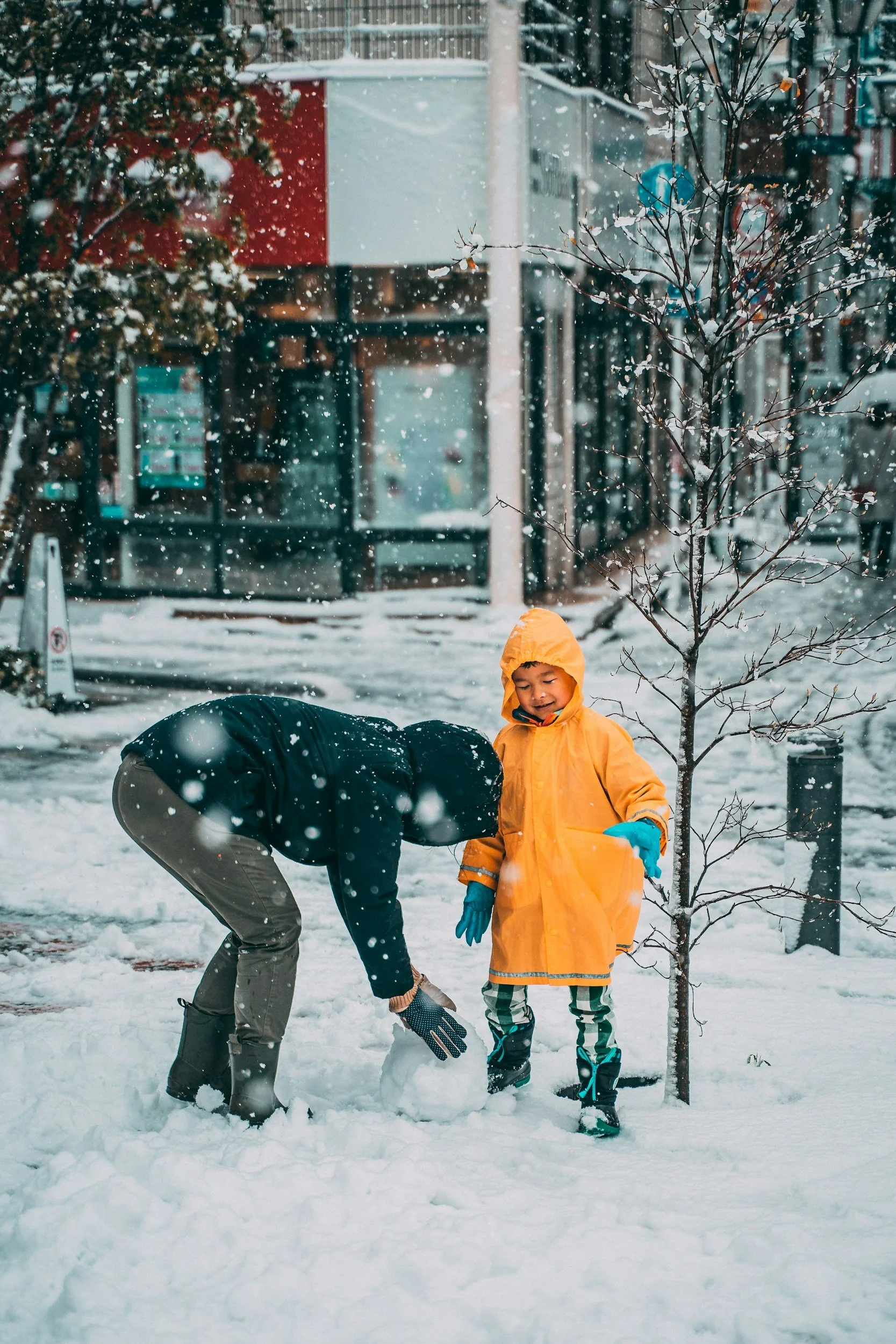 A man building a snowman with a young child in a bright yellow coat on a snowy day, both focused on the moment without any devices in sight.