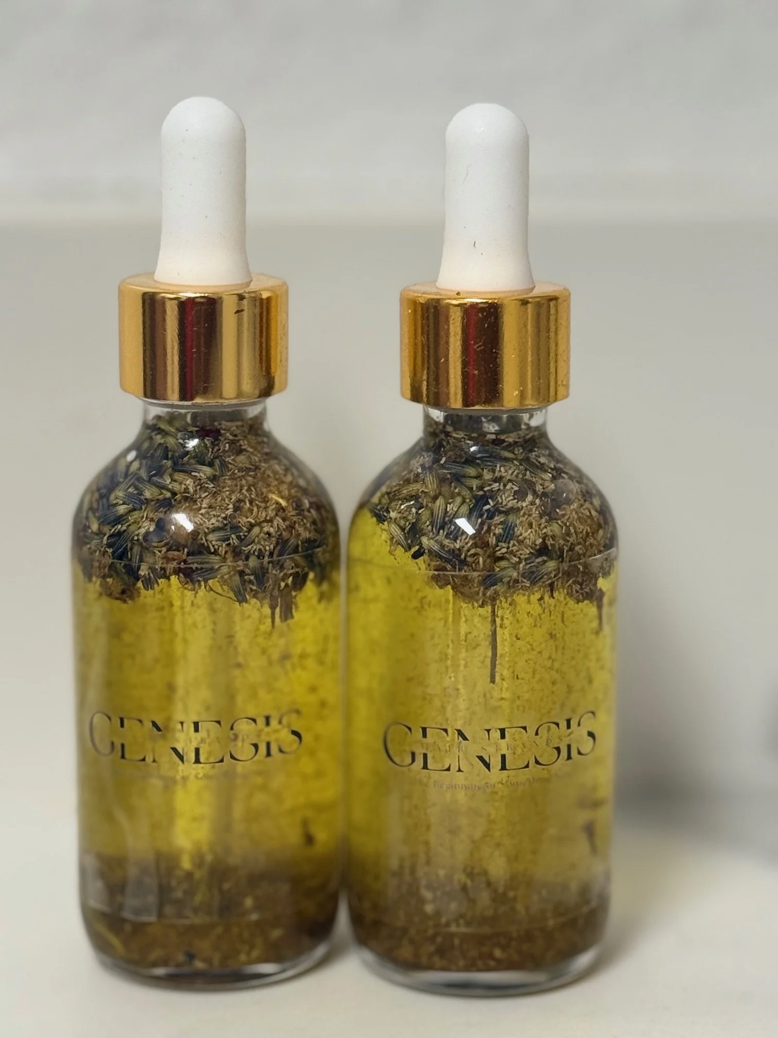 Two glass dropper bottles labeled 'GENSEIS' with dried herbs or flowers inside, capped with white droppers and gold rings, placed against a plain background.