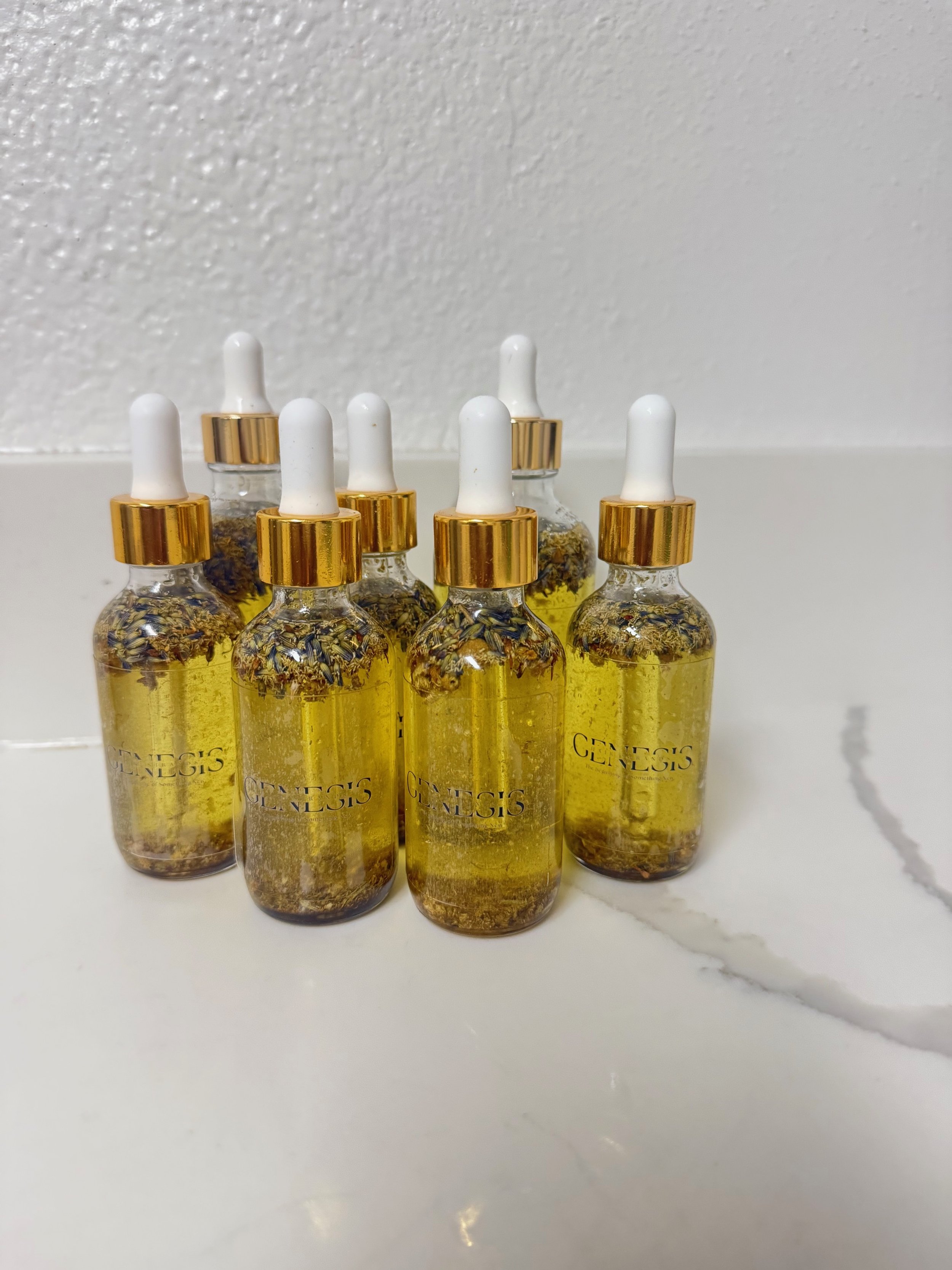 Six small glass bottles with white droppers and gold caps, labeled 'GENESIS', filled with a yellow herbal infusion with dried herbs inside, on a white surface with a white textured background.