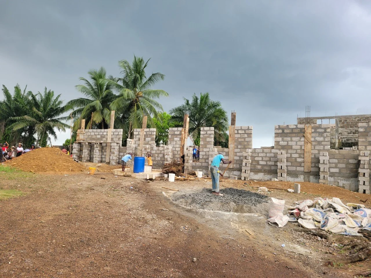 Construction site with workers building a brick wall, piles of dirt and construction materials, tall palm trees in the background, and a cloudy sky overhead.